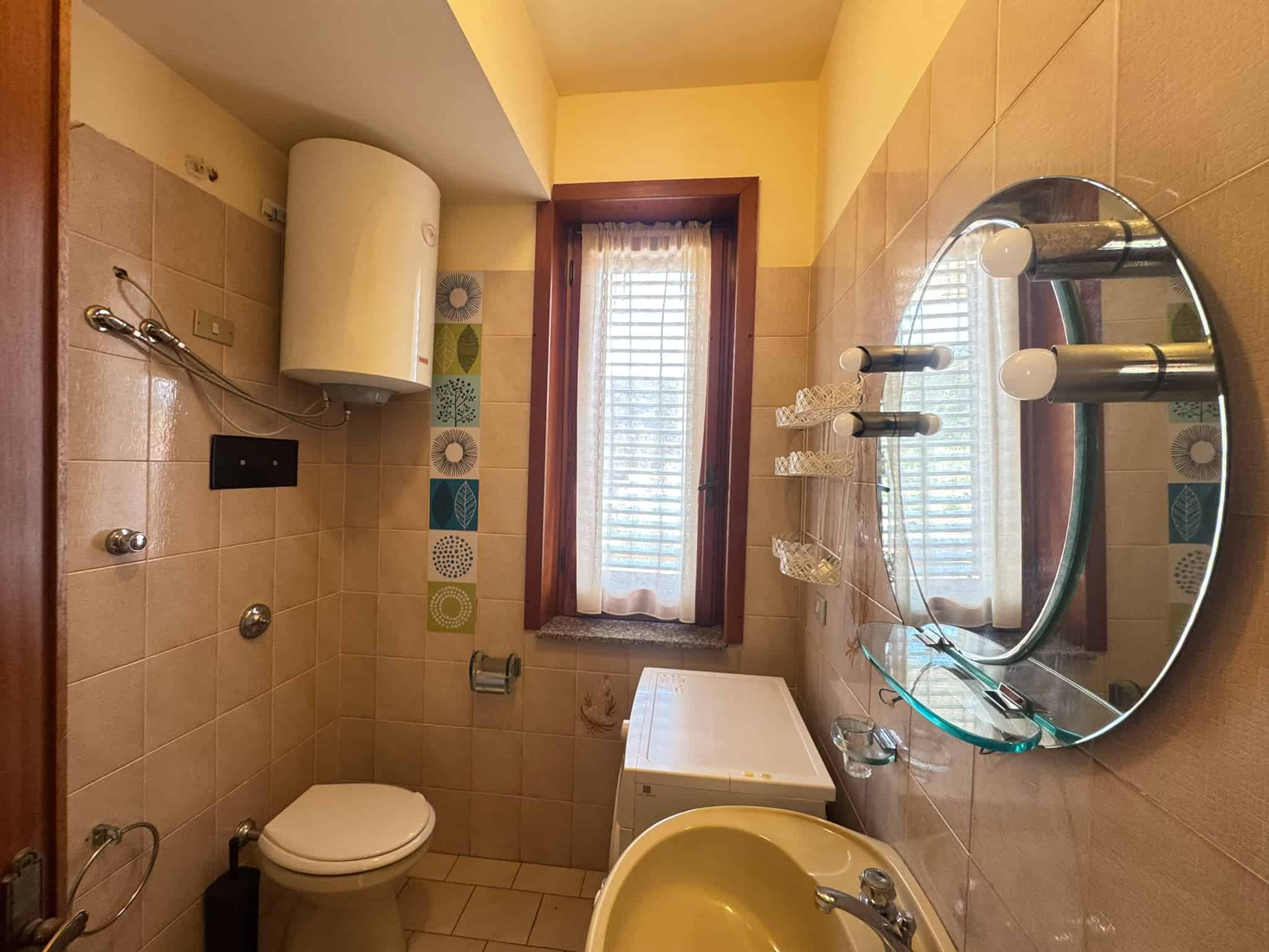 Bagno con pareti rivestite in piastrelle beige, lavabo con specchio rotondo e finestra con tende in tessuto.