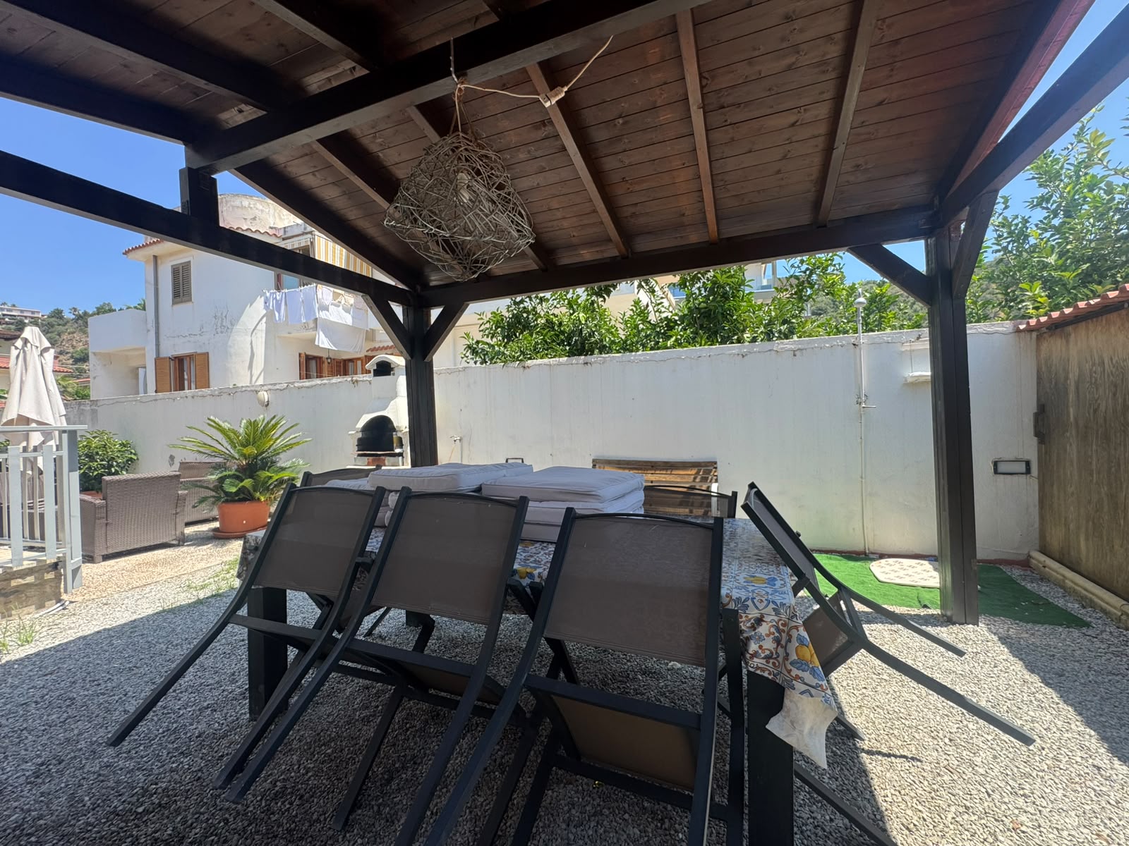 Zona esterna con tavolo e sedie sotto un pergolato, giardino con piante e area relax.