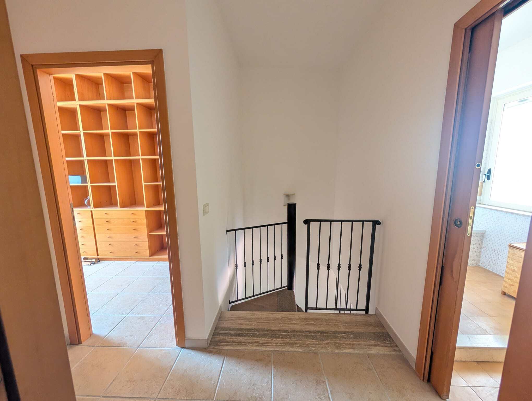 Ingresso di una villetta a schiera con scale in legno e corrimano in ferro, vista da un corridoio interno con porte in legno e una stanza luminosa.