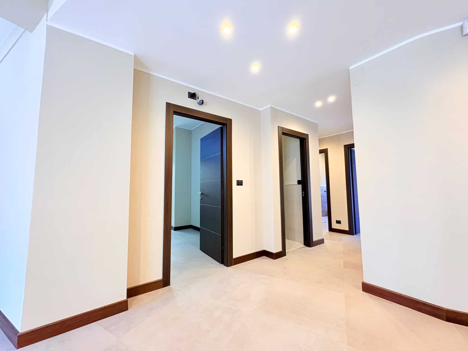 Interno di un appartamento moderno con pareti bianche e porte in legno scuro, illuminato da luci a soffitto, in zona residenziale a Milazzo.