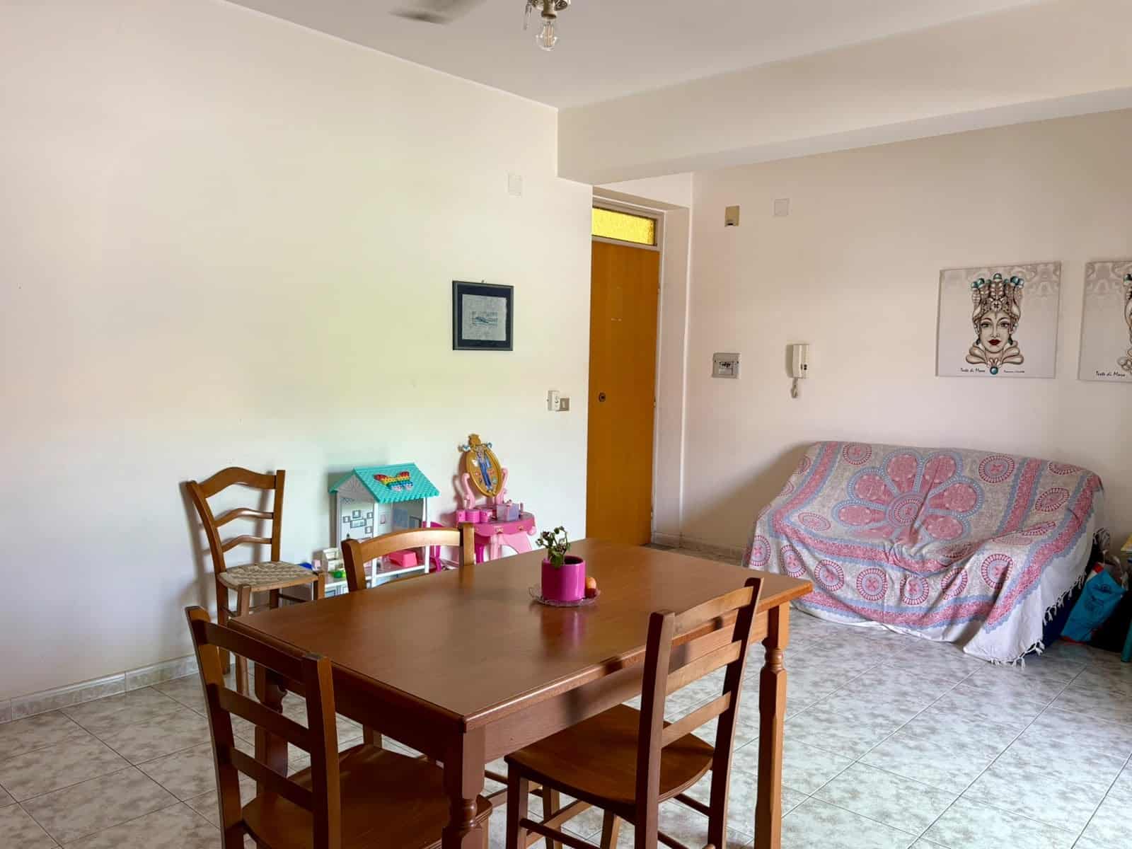 Interno di un appartamento con tavolo in legno, divano coperto da una coperta colorata e pareti decorate con quadri, in zona soggiorno.