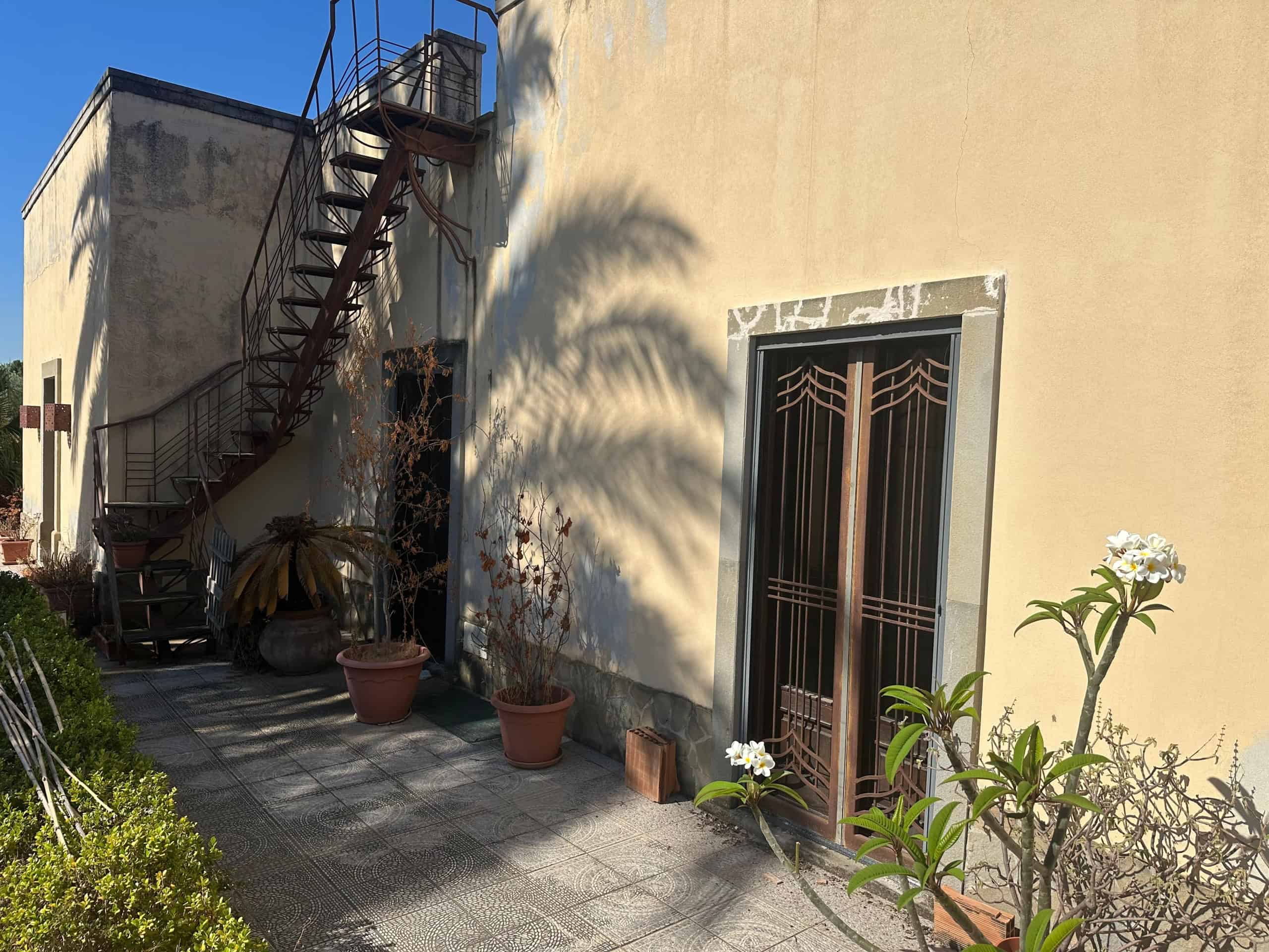 Facciata di una villa con porta in ferro battuto, piante in vaso e scala esterna in metallo, ombra di palma sulla parete gialla.