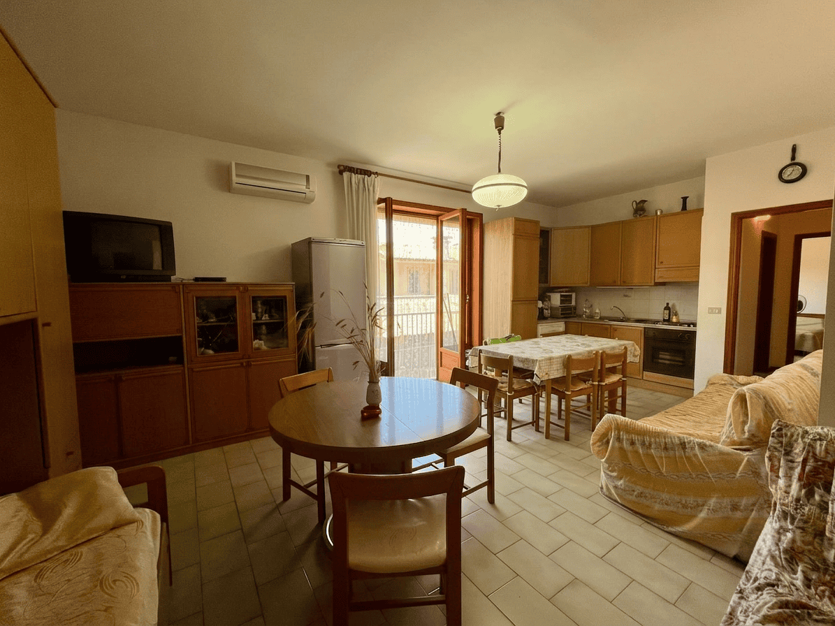 Interno di un appartamento con soggiorno e cucina, arredato con mobili in legno e divani, finestra con balcone e illuminazione centrale.