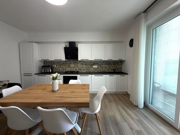 Cucina moderna con mobili bianchi, tavolo in legno e sedie bianche, grande finestra con tende trasparenti, ideale per appartamento in vendita a Milazzo.