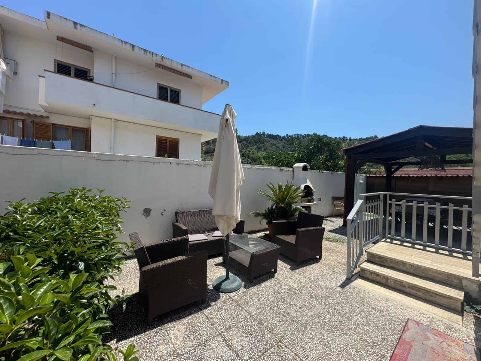 Spazio esterno di una villa in vendita a Milazzo con area relax, sedie, tavolo e ombrellone sotto un cielo sereno.