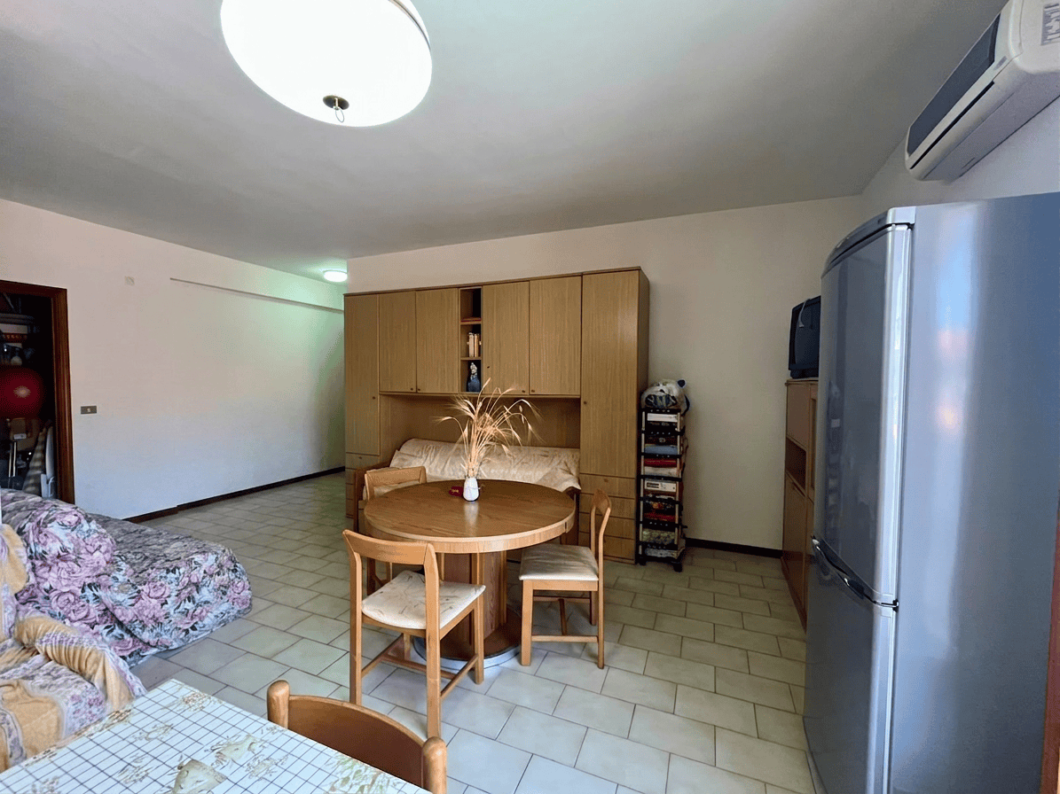 Interno di un appartamento con soggiorno, tavolo rotondo, sedie, cucina e frigorifero in stile semplice e funzionale.