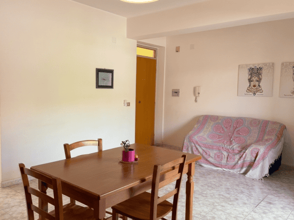 Interno di un appartamento con tavolo in legno e divano con copertura colorata, in vendita in via S. Maria 80, Tonnarella, Furnari, Messina.