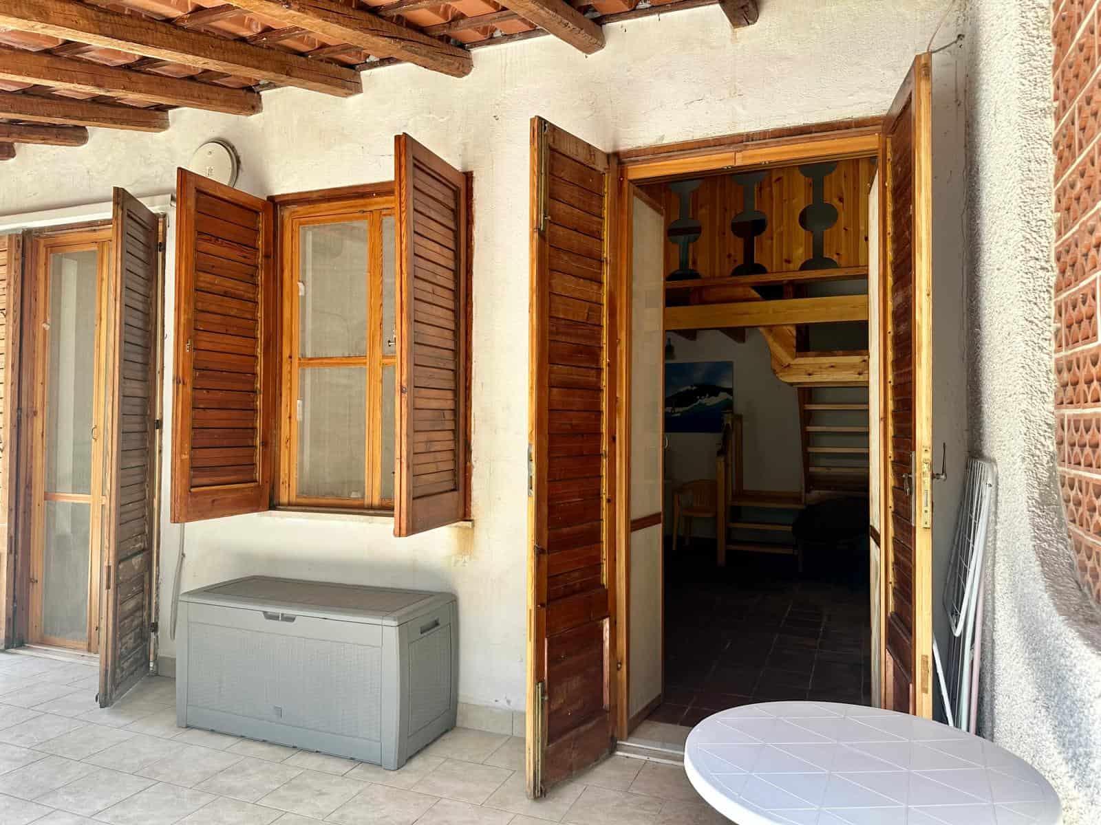 Finestra in legno e porta aperta che conduce a un interno con scala in legno, in un ambiente esterno con pareti in pietra e terrazza.