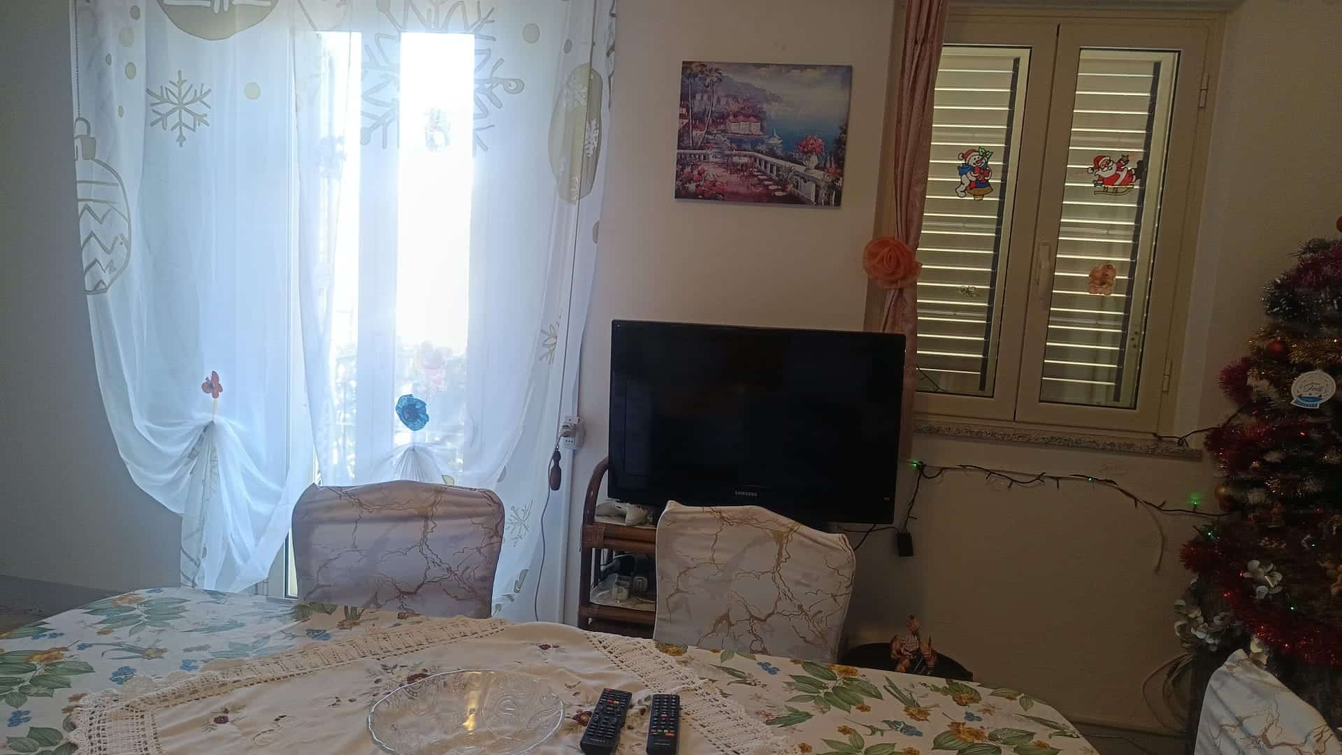 Interno di un appartamento con soggiorno, TV, decorazioni natalizie e finestra con tende leggere.