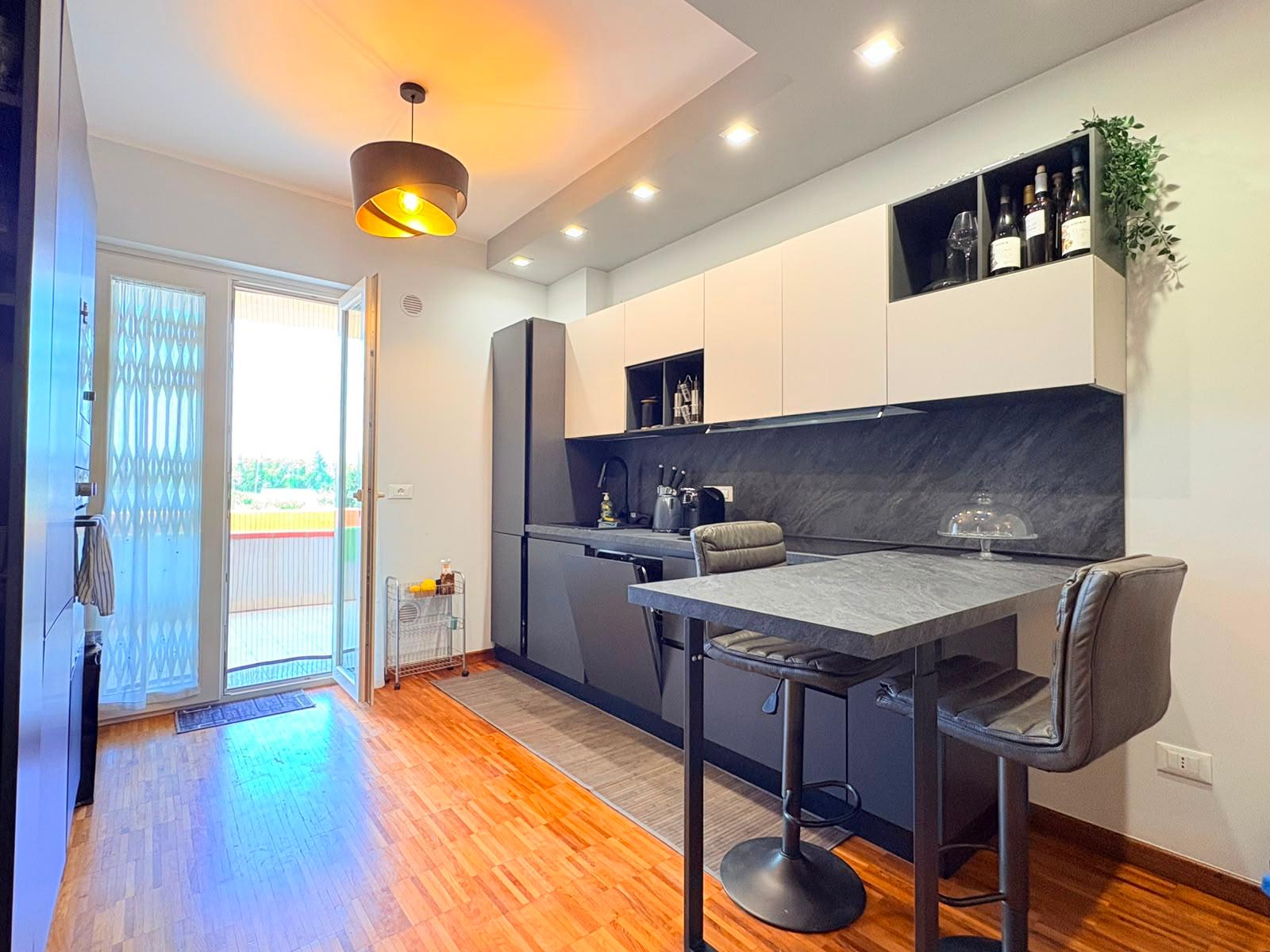 Cucina moderna con mobili bianchi e parete in marmo nero, balcone con vista aperta, illuminazione a soffitto e tavolo con sedie in soggiorno.