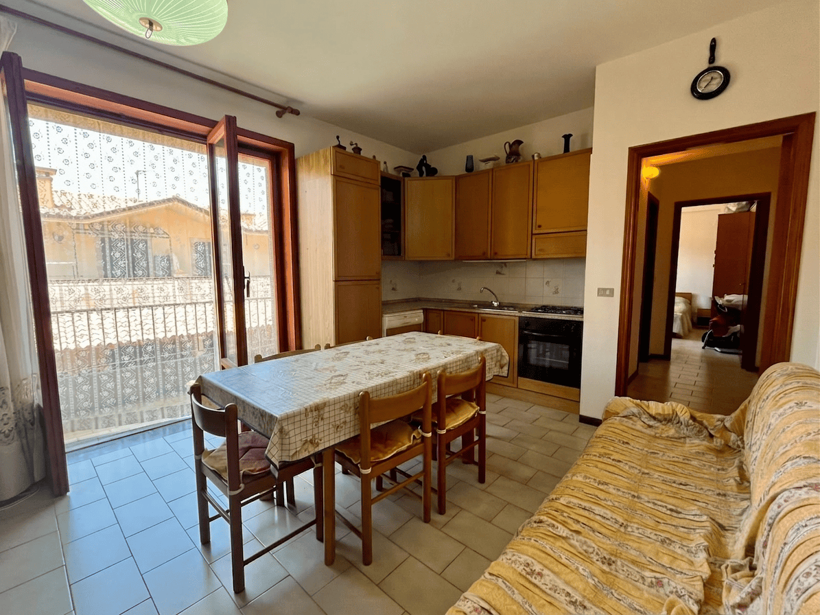 Interno di un appartamento con cucina e zona pranzo, vista su balcone e soggiorno.