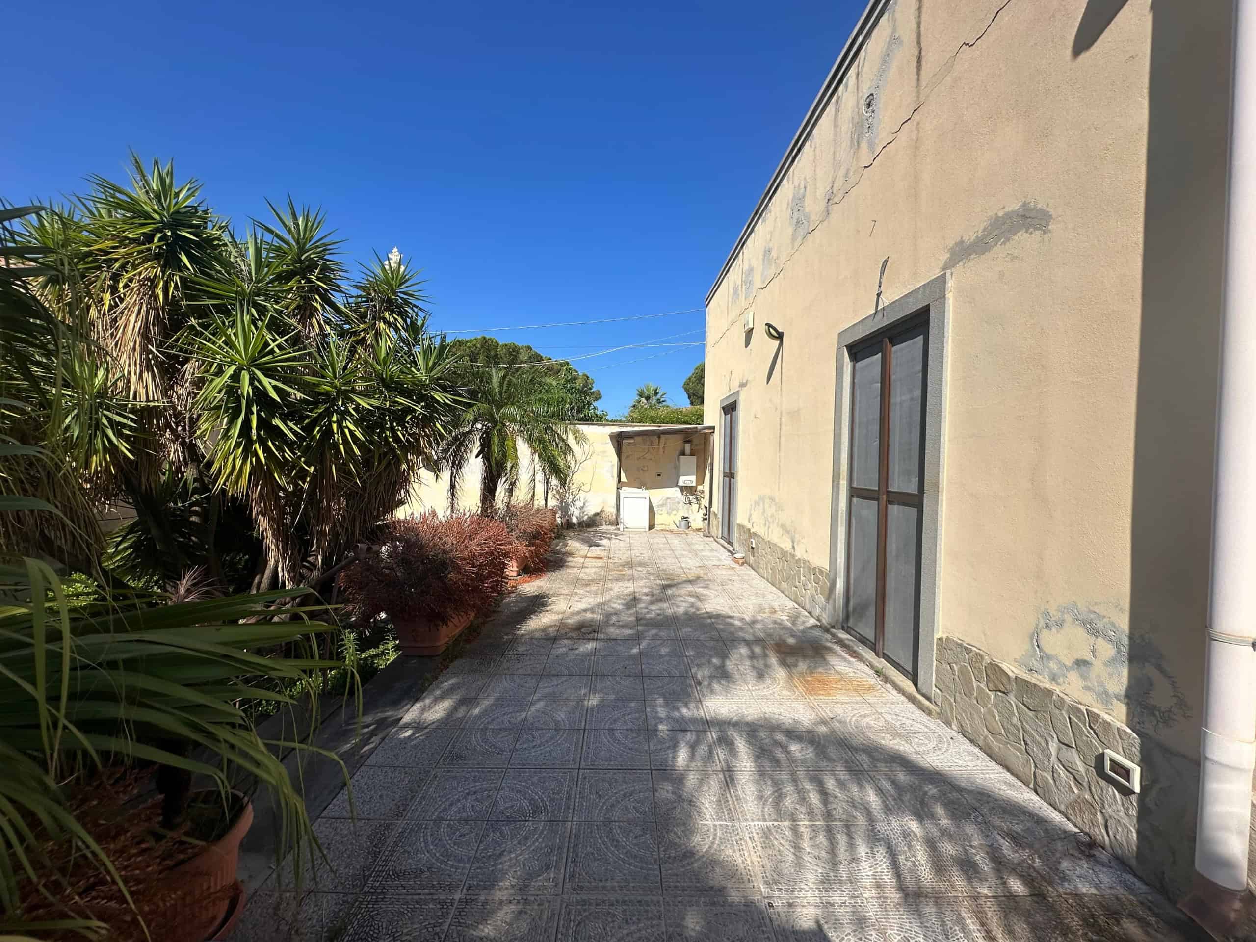Vicolo laterale di una villa in vendita a Milazzo con piante tropicali e pareti beige sotto un cielo azzurro.
