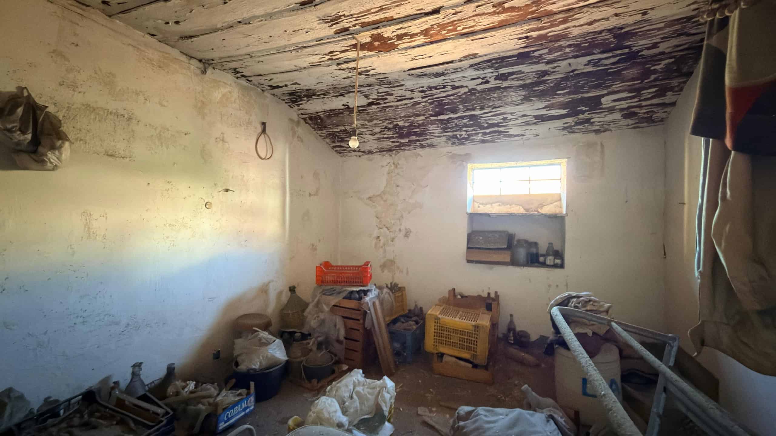 Interno di una stanza abbandonata con pareti danneggiate e oggetti disordinati, luce naturale dalla finestra.
