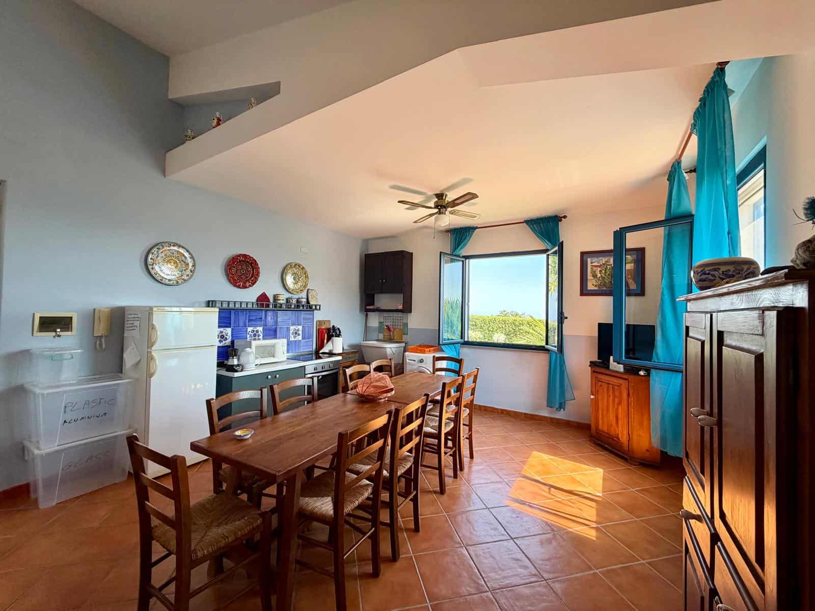Interno di una sala da pranzo luminosa con tavolo in legno, sedie, grandi finestre con tende azzurre e vista panoramica.