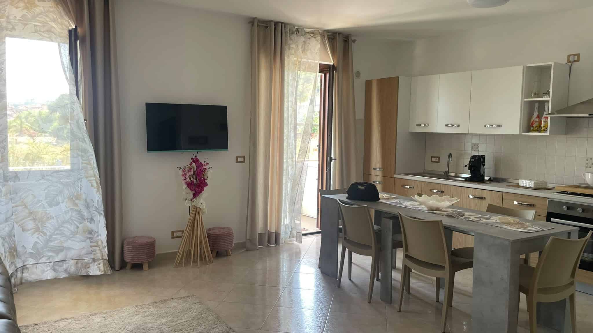 Interno di un appartamento con soggiorno e cucina moderna, grande finestra e porta finestra che dà sul balcone, arredamento semplice e funzionale.