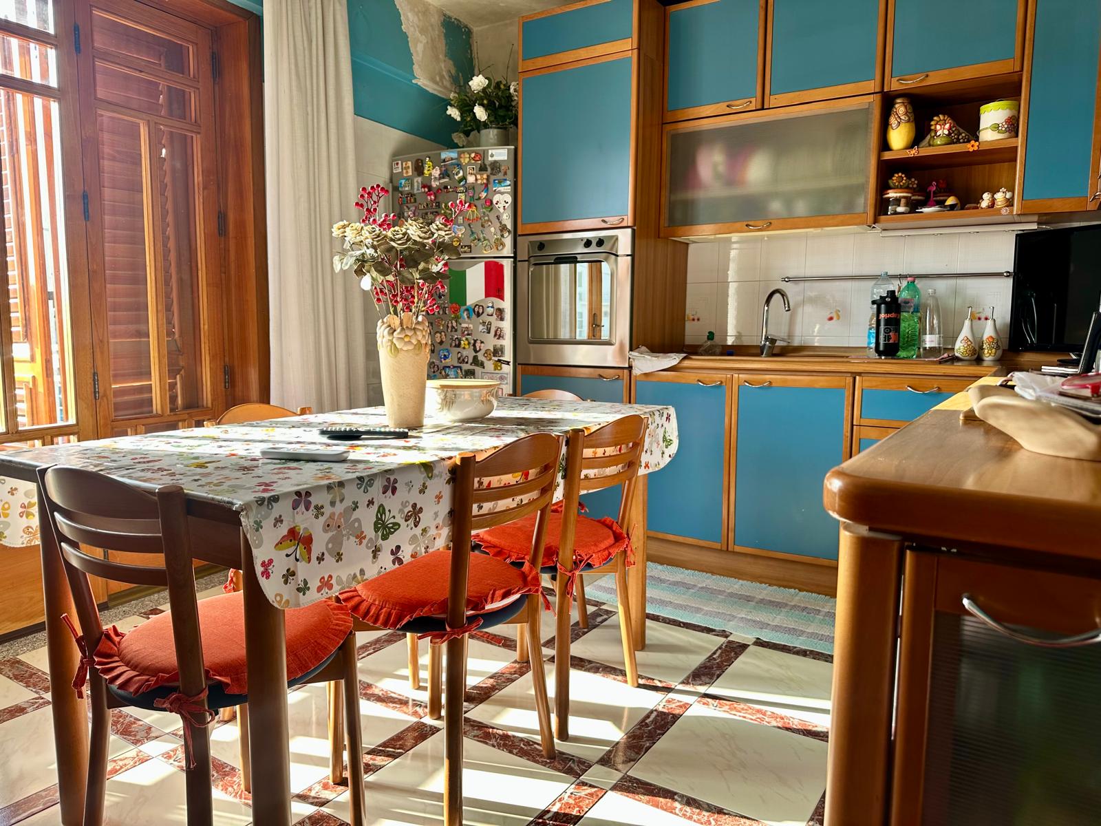 Cucina abitabile con mobili blu e legno, tavolo con tovaglia floreale e sedie con cuscini rossi, ambiente luminoso con grande finestra e decorazioni colorate.