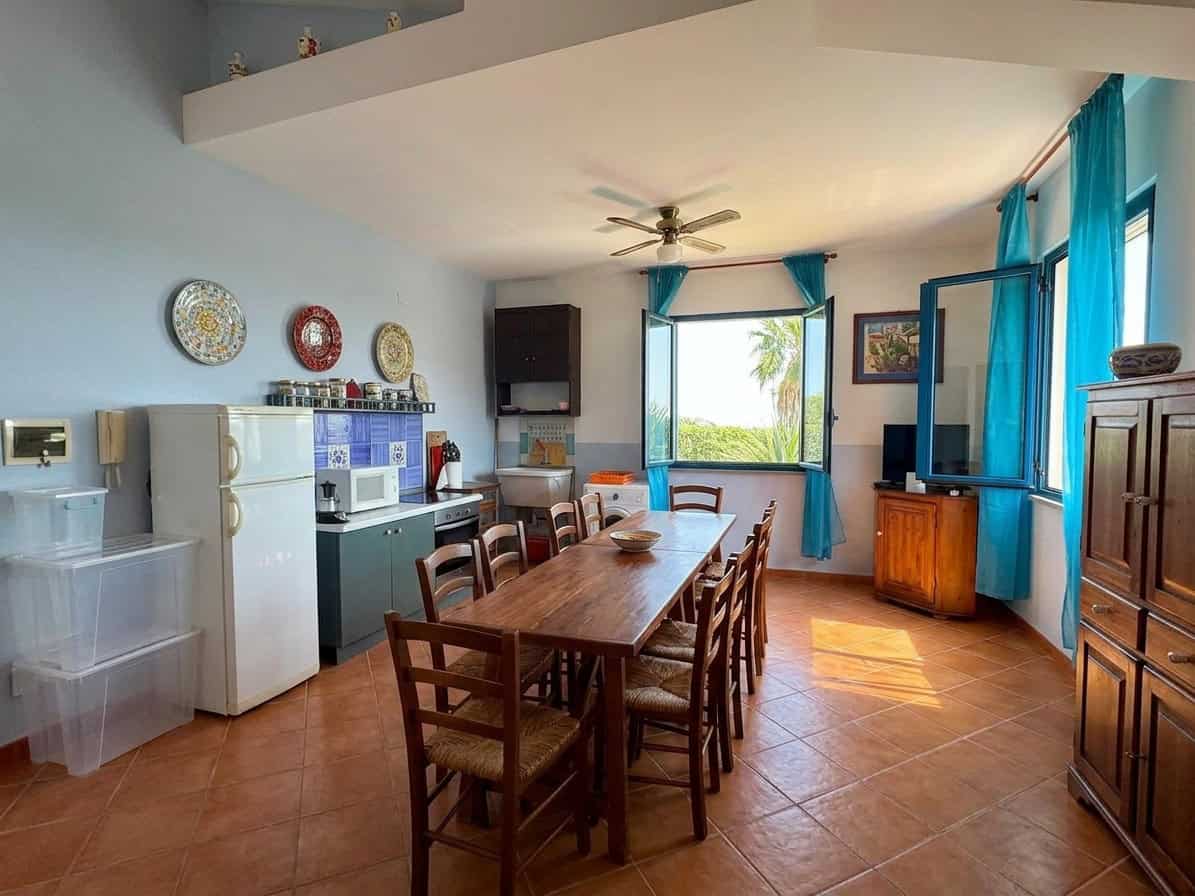 Interno di una sala da pranzo luminosa con tavolo in legno, sedie, grandi finestre con tende azzurre e vista sul giardino tropicale.