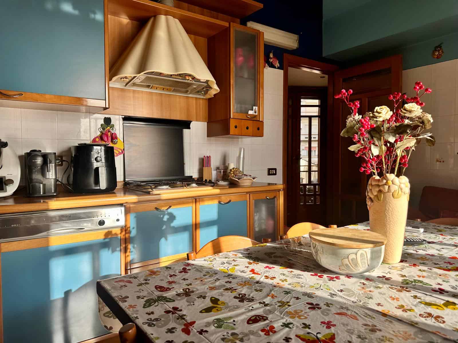 Cucina moderna con mobili in legno e pareti blu, tavolo con tovaglia floreale e vaso di fiori colorati, ambiente accogliente e luminoso.