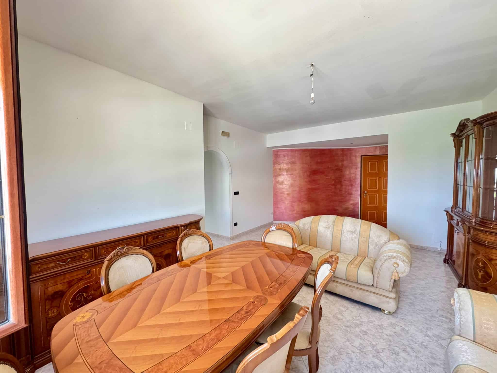 Interni di un soggiorno spazioso con mobili in legno e divani in tessuto beige, pareti bianche e una parete rossa, ambiente luminoso e accogliente.