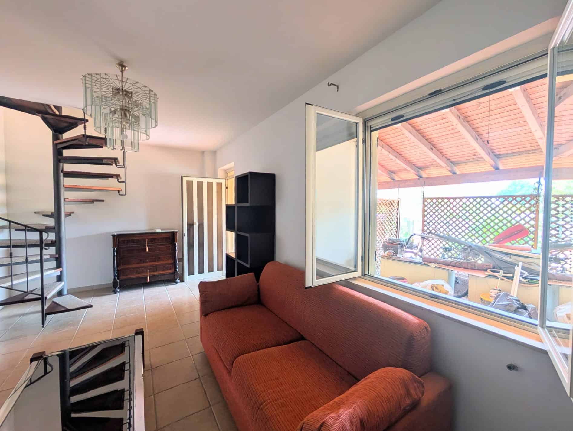Interno di una villetta a schiera con divano rosso, scala a spirale e vista sul patio esterno.