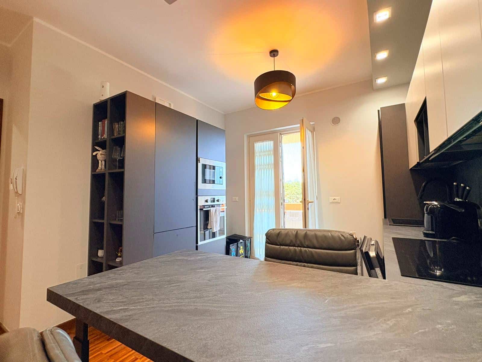 Cucina moderna con armadi grigi, forno e microonde integrati, tavolo in pietra e sedie nere, illuminazione soffusa e porta finestra che dà sul balcone.