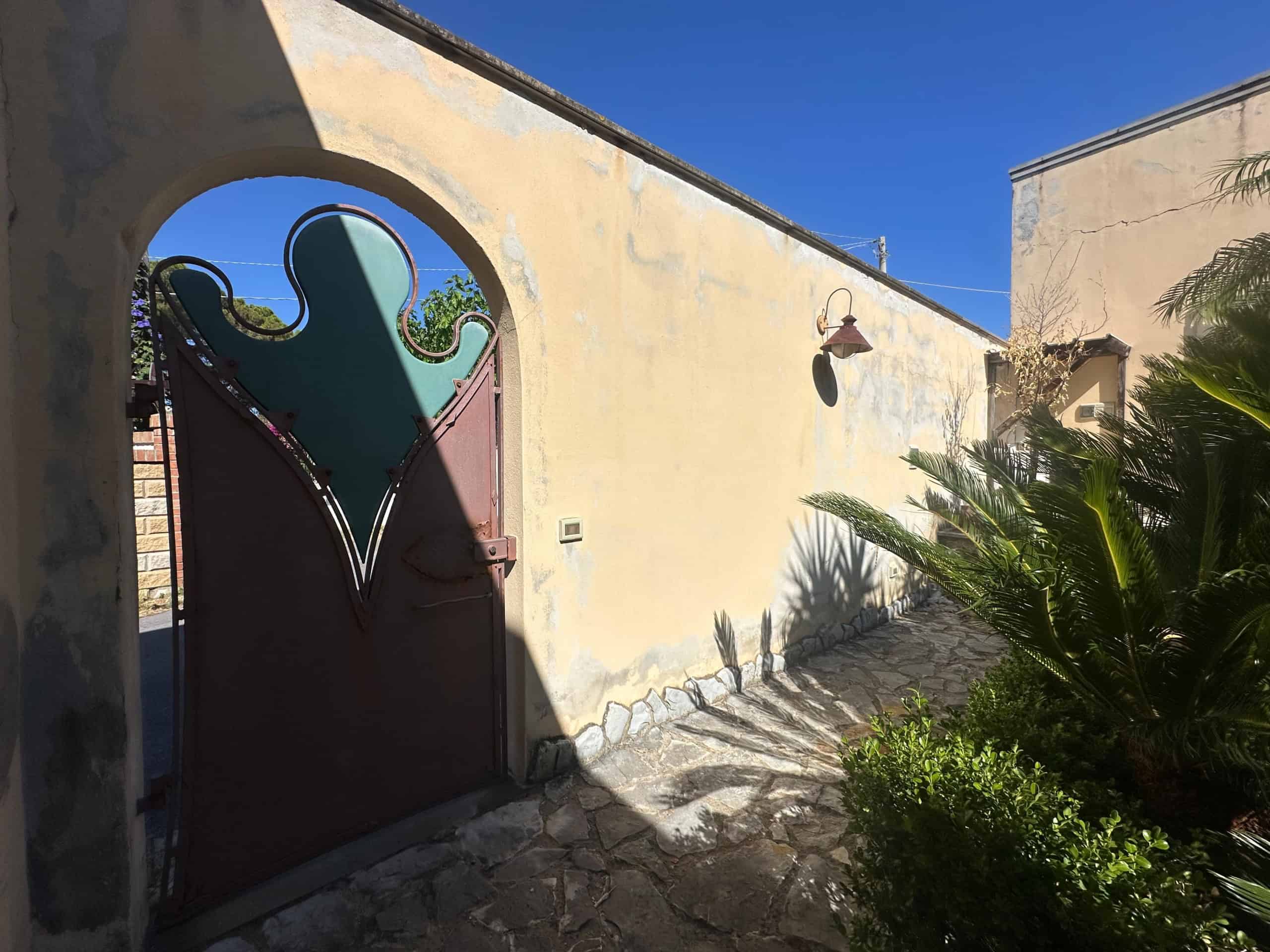 Porta in ferro battuto con decorazioni artistiche, ingresso a una proprietà a Milazzo, vista sotto un cielo azzurro.
