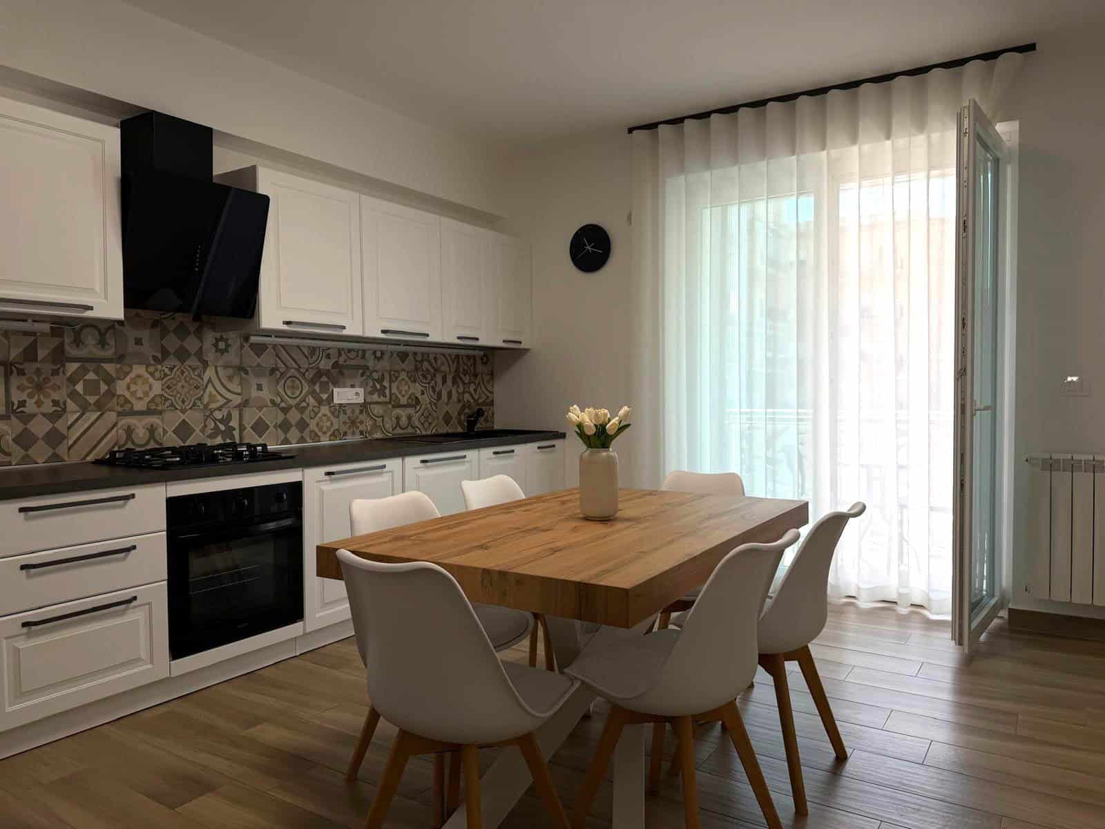 Cucina moderna con tavolo in legno e sedie bianche, grande finestra con tende trasparenti, pareti bianche e pavimento in legno chiaro.