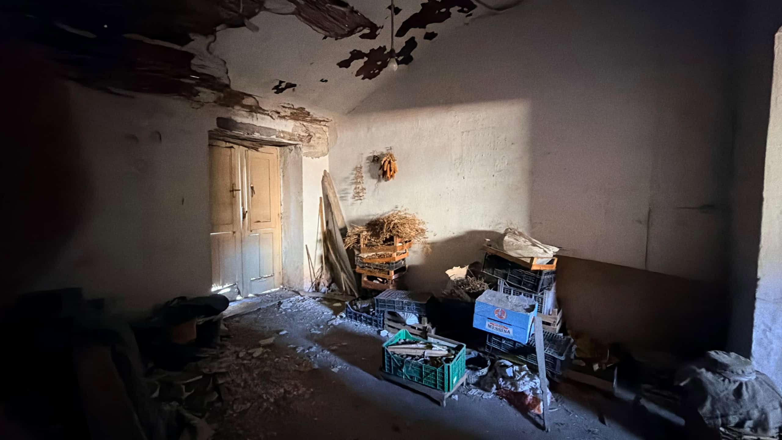 Interno di una stanza abbandonata con pareti danneggiate e oggetti disordinati, evidenziando condizioni di degrado e abbandono.