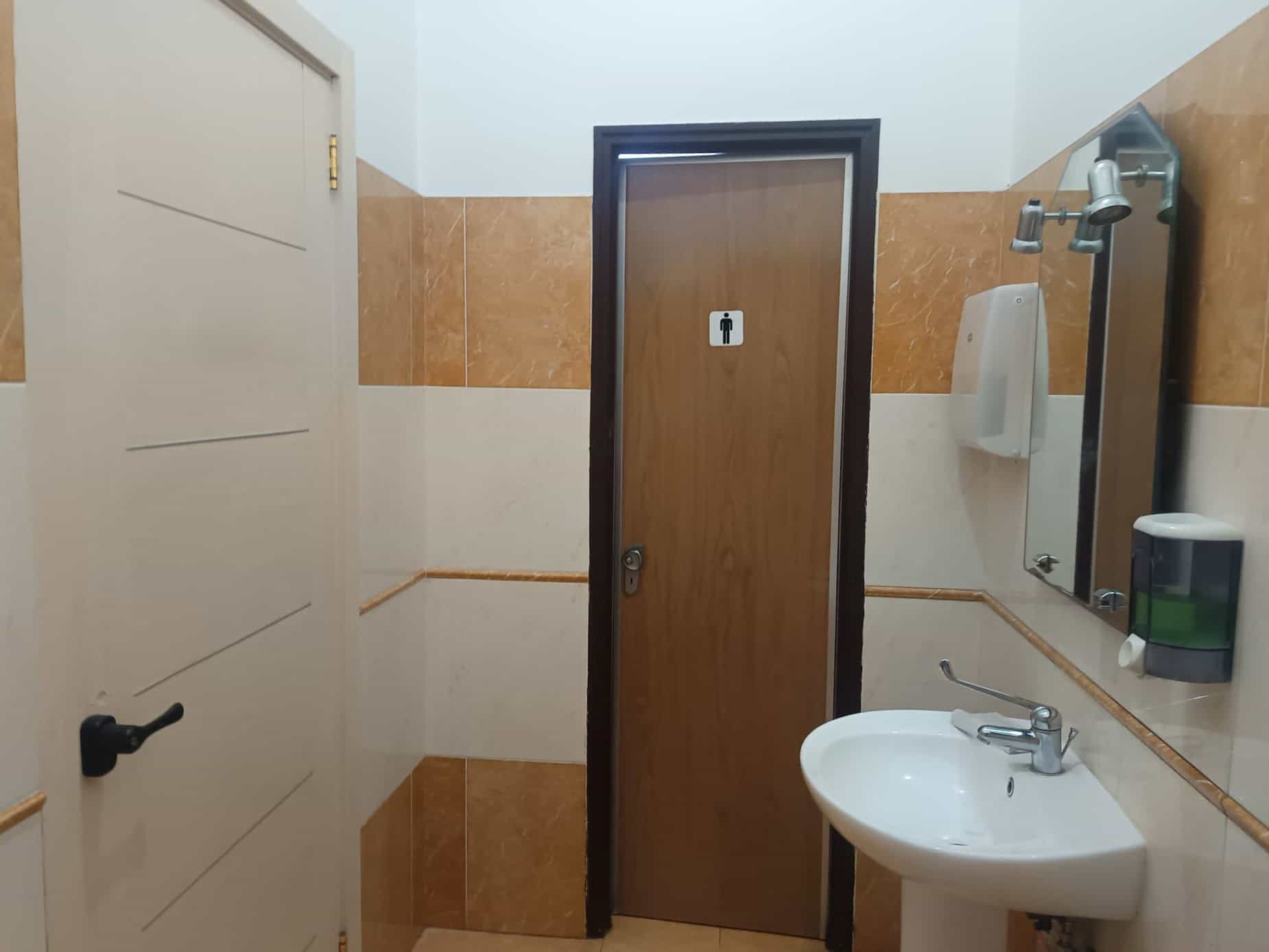 Bagno pubblico con lavabo, specchio e dispenser di sapone, in un ambiente con pareti rivestite in piastrelle beige e marroni.