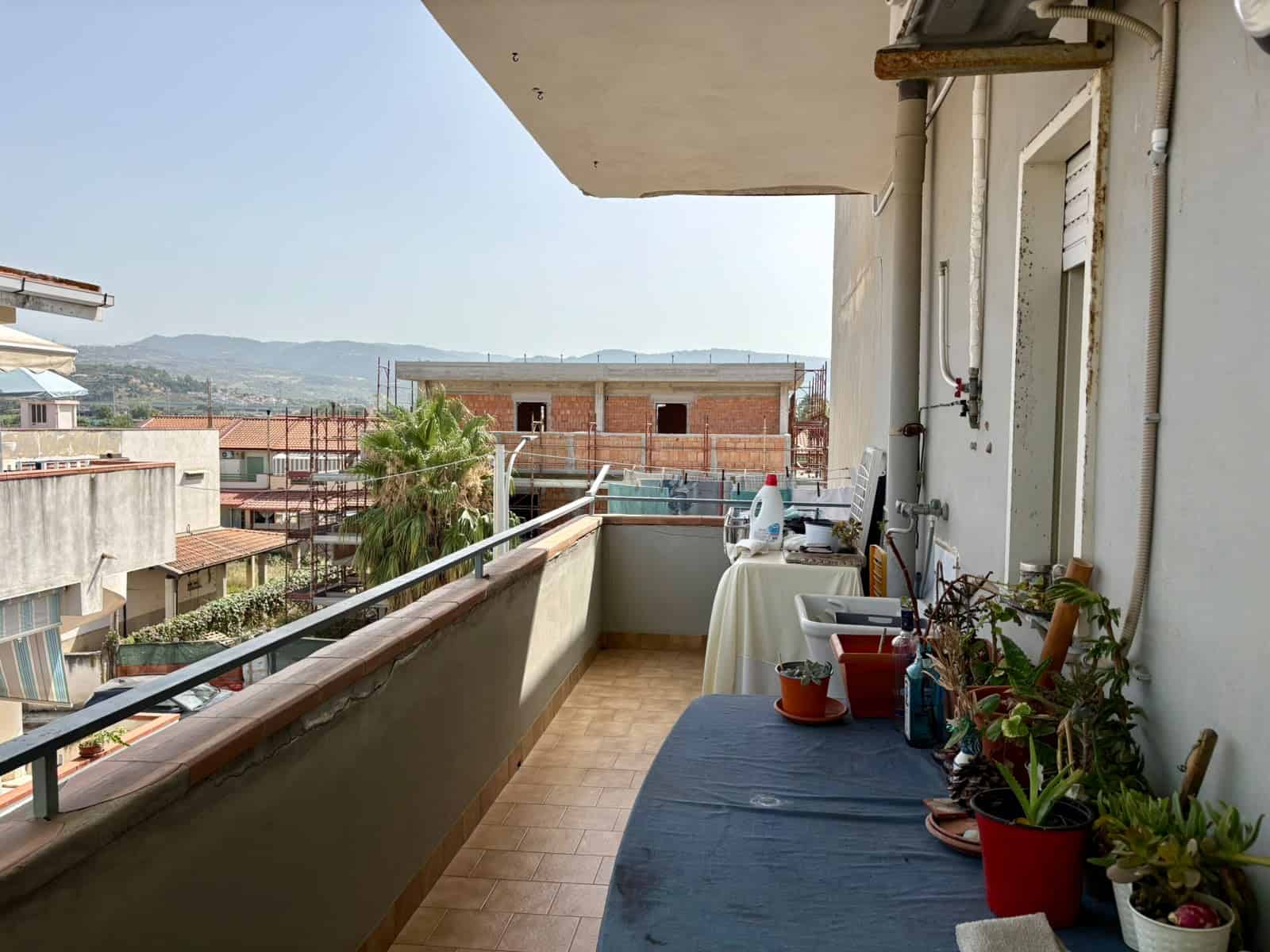Balcone di un appartamento con vista su edifici e montagne, decorato con piante e oggetti vari.