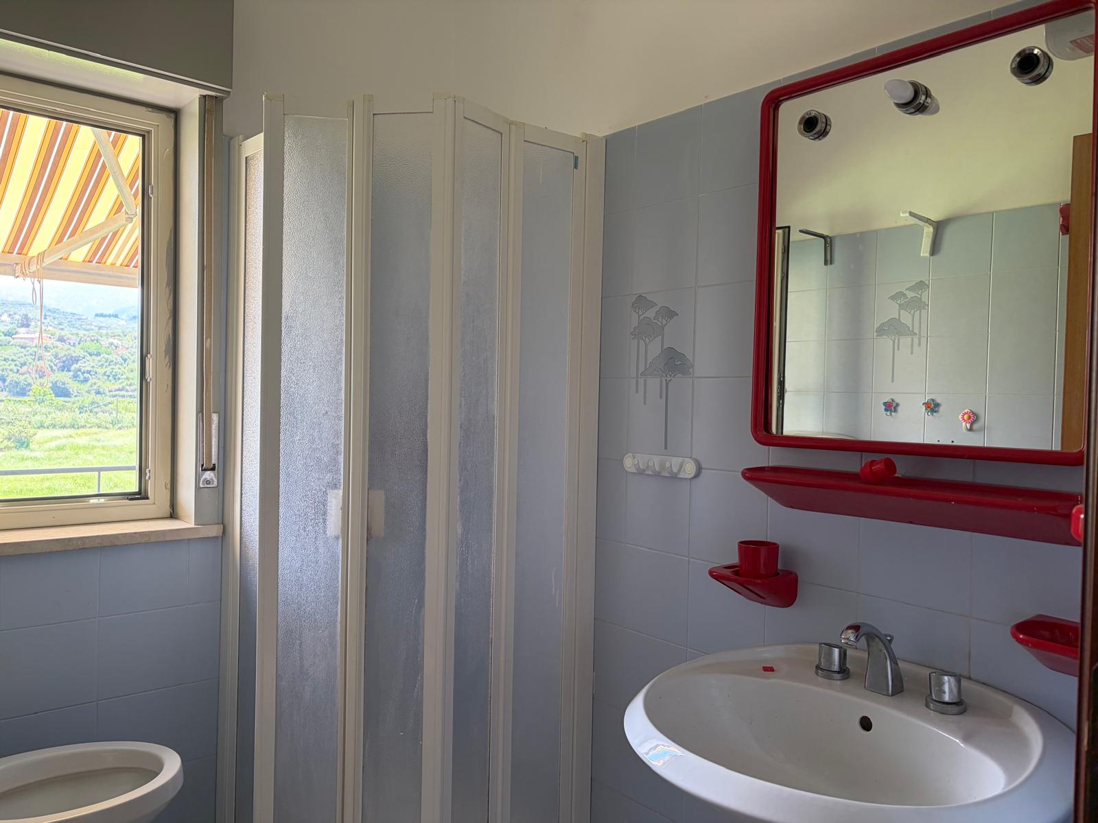 Bagno moderno con specchio rosso, lavabo e finestra con vista panoramica sulla campagna.