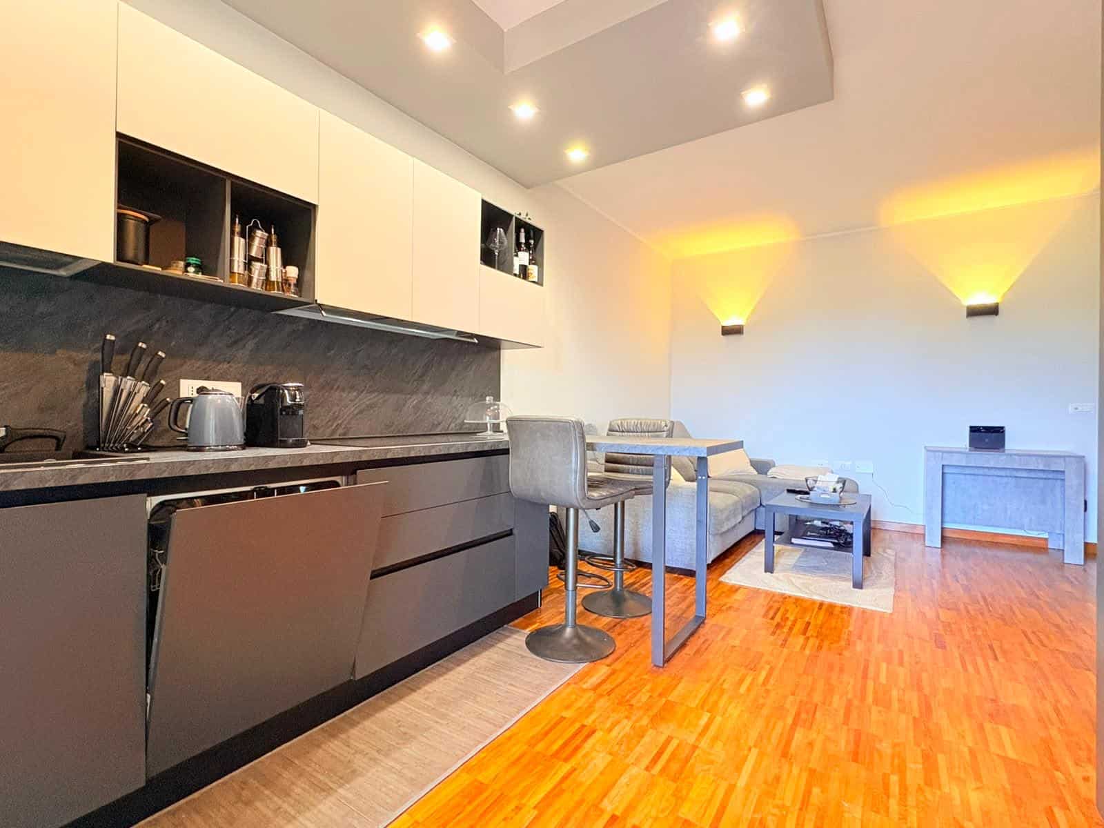 Interno di un appartamento moderno con cucina a vista e zona soggiorno con divano e tavolino, illuminazione calda e pavimento in legno.