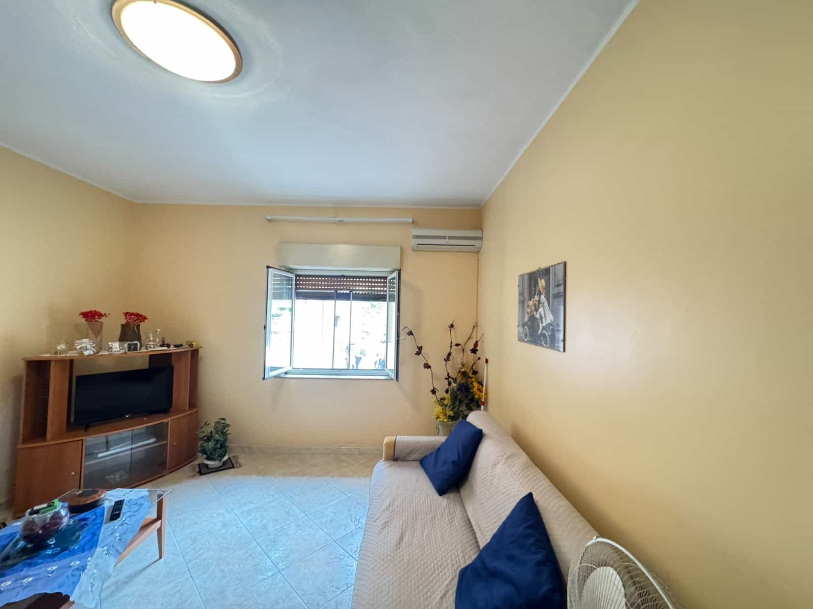Interno di un soggiorno luminoso con pareti gialle, divano beige con cuscini blu, finestra con vista esterna e mobile TV in legno.