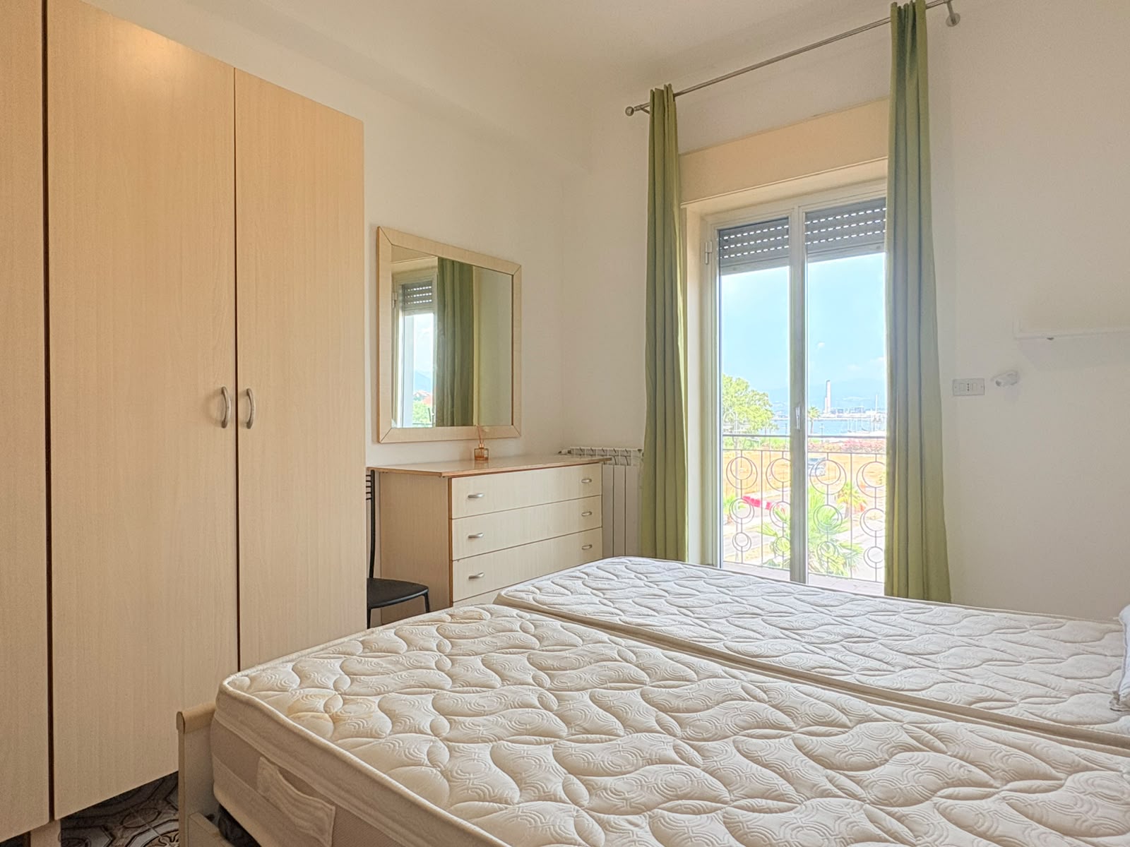Camera che mostra una camera da letto luminosa con armadio, cassettiera, specchio e balcone con vista sul mare a Milazzo.