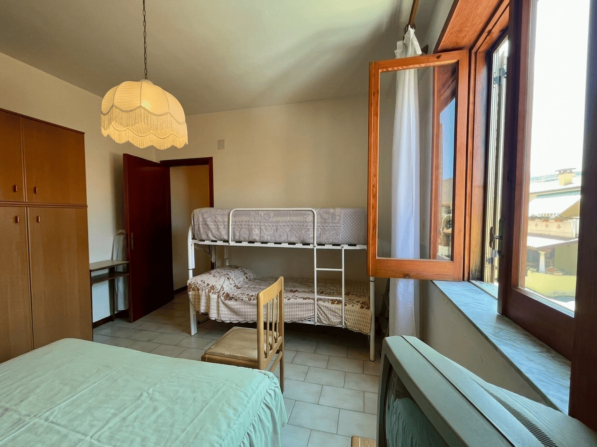 Camera che mostra una stanza con letto matrimoniale, letto a castello, armadio e finestra aperta con vista su case e cielo.