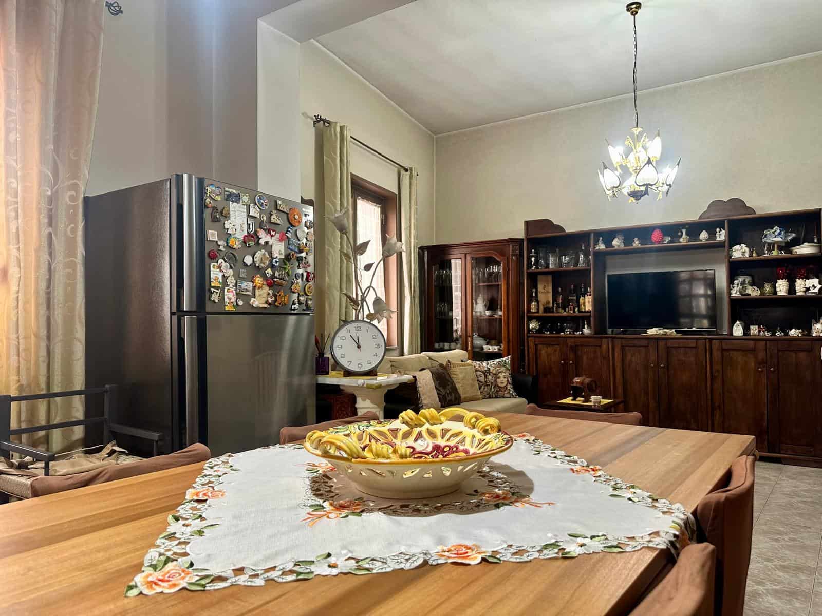 Interno di una cucina con frigorifero, tavolo e mobile con TV e oggetti decorativi, ambiente accogliente e ben illuminato.