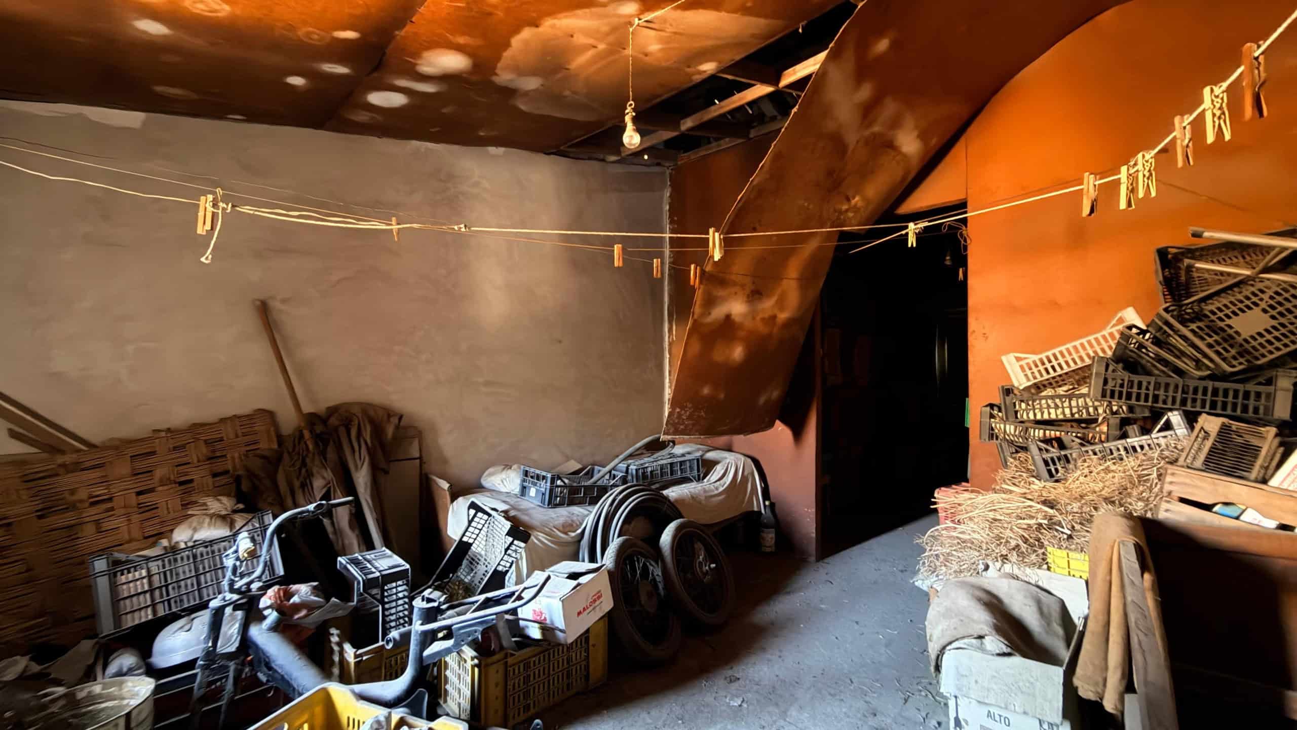 Interno di un garage disordinato con attrezzi, scatole e materiali vari, pareti e soffitto danneggiati.
