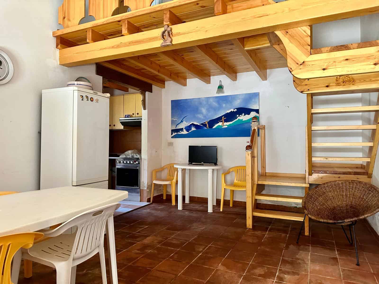 Interno di un appartamento con cucina e zona soggiorno, soffitto in legno e scala in legno che porta al soppalco, arredamento semplice e funzionale.
