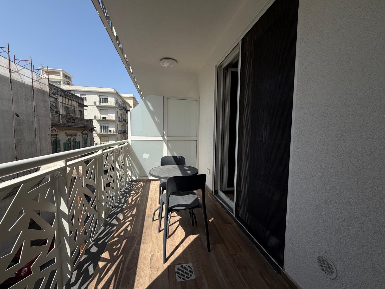 Balcone di un appartamento a Milazzo con sedie e vista su edifici circostanti, pavimento in legno e ringhiera decorativa, in una giornata soleggiata.