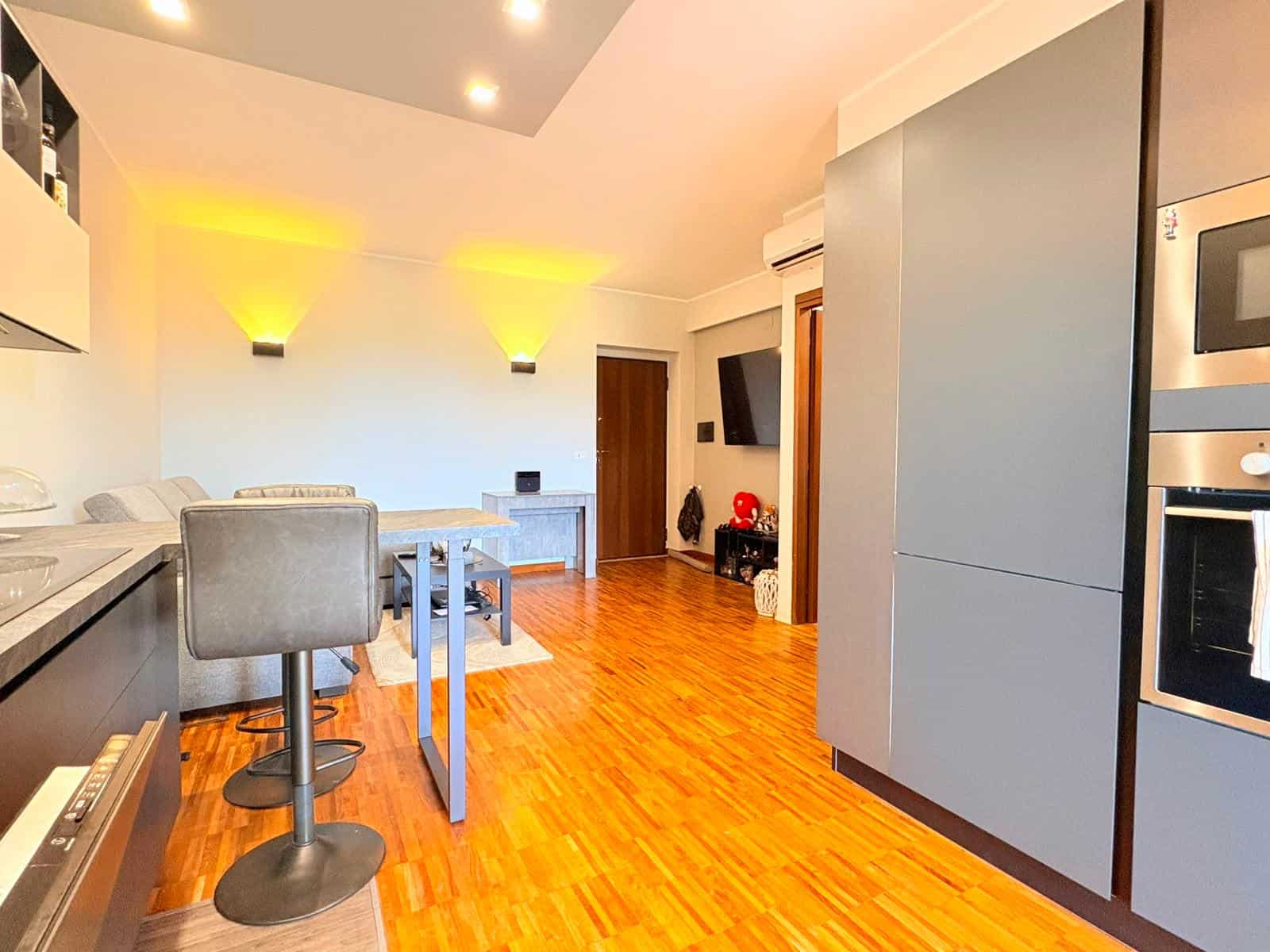 Interno di un appartamento moderno con cucina e zona soggiorno, pavimento in legno e illuminazione soffusa.