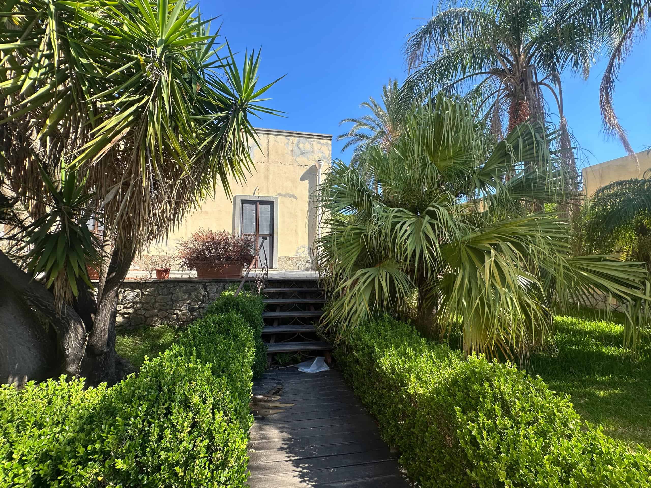 Giardino con piante tropicali e sentiero in pietra, vista di una villa in vendita a Milazzo, ME.