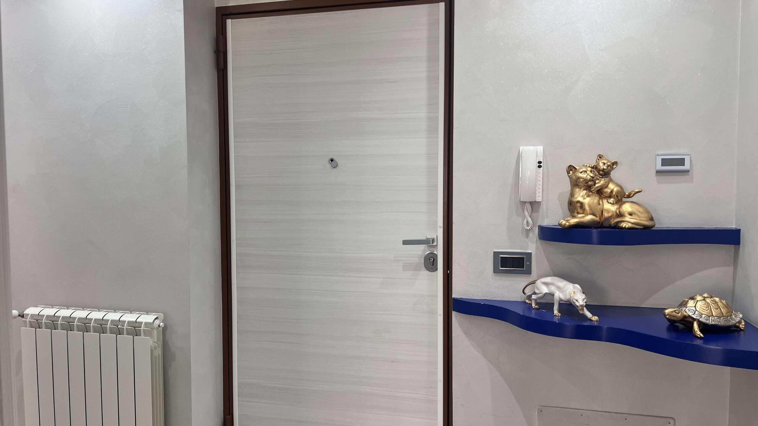 Ingresso con porta bianca e decorazioni eleganti, tra cui un gatto dorato e una tartaruga su mensole blu, in un ambiente luminoso e accogliente.