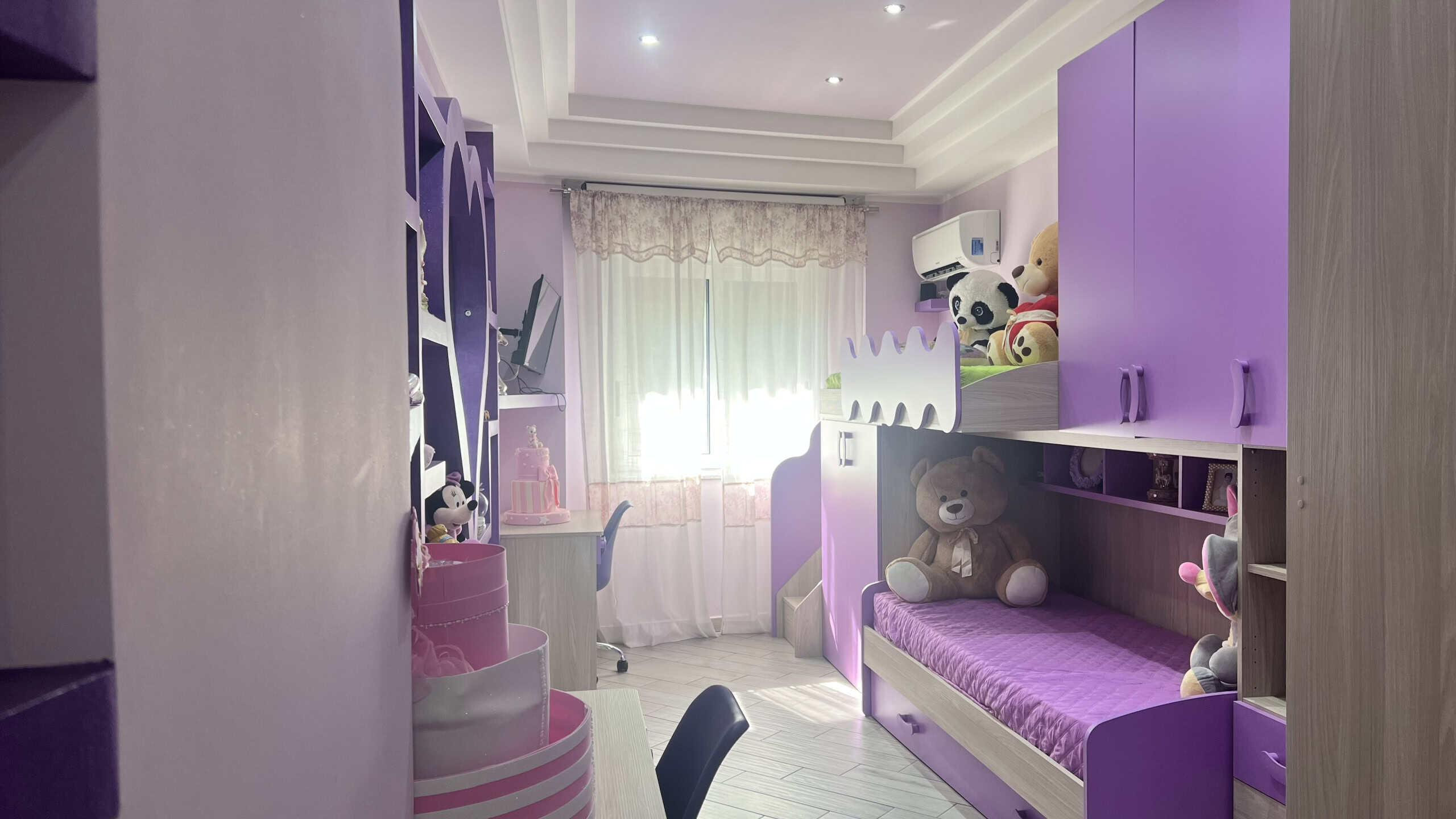 Interno di una camera da letto moderna con mobili viola e decorazioni per bambini, luce naturale dalla finestra, ambiente accogliente e colorato.