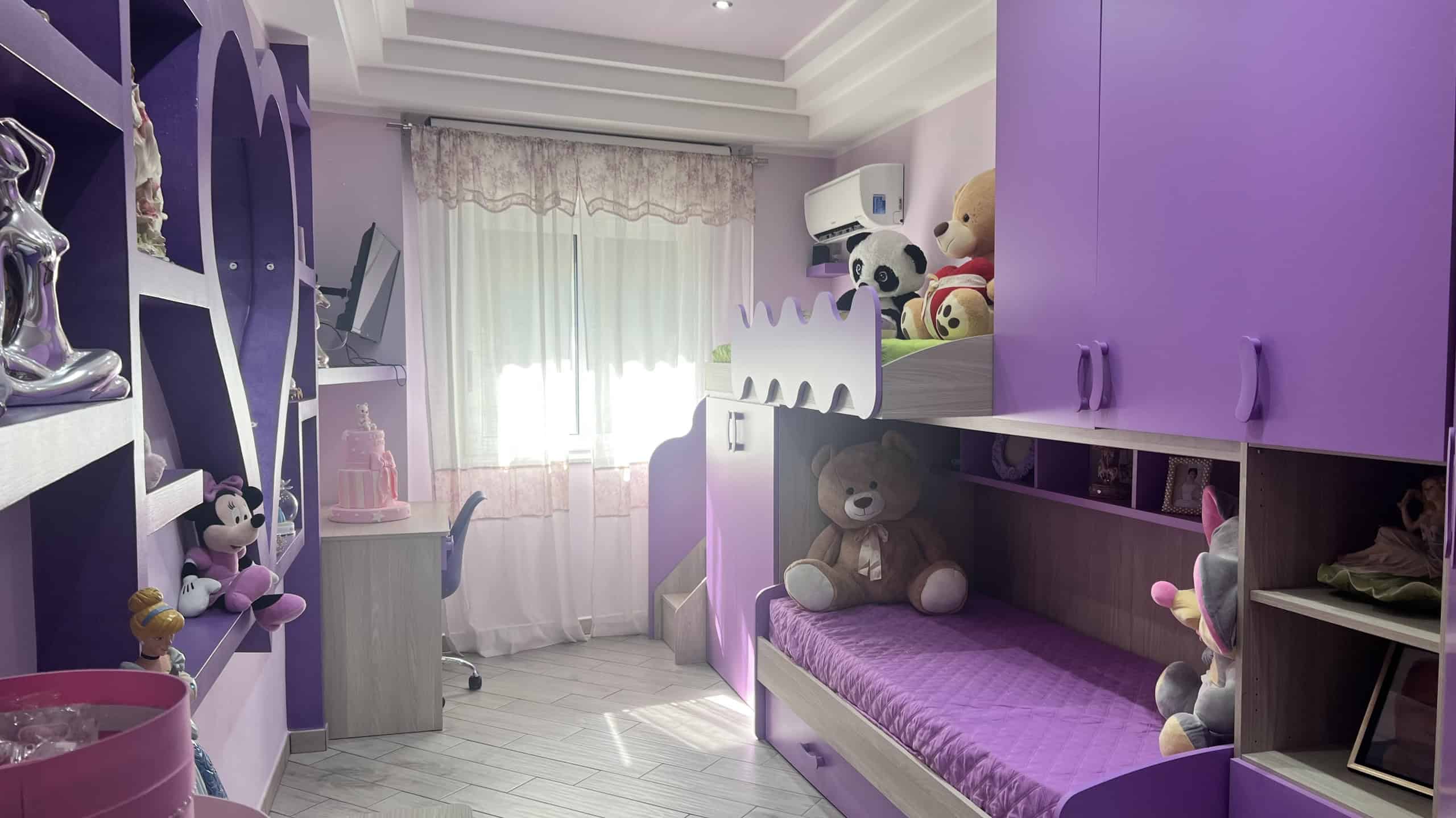 Interno di una camera da letto color lavanda con armadio, scrivania e numerosi peluche, luminosa grazie alla finestra con tende leggere.