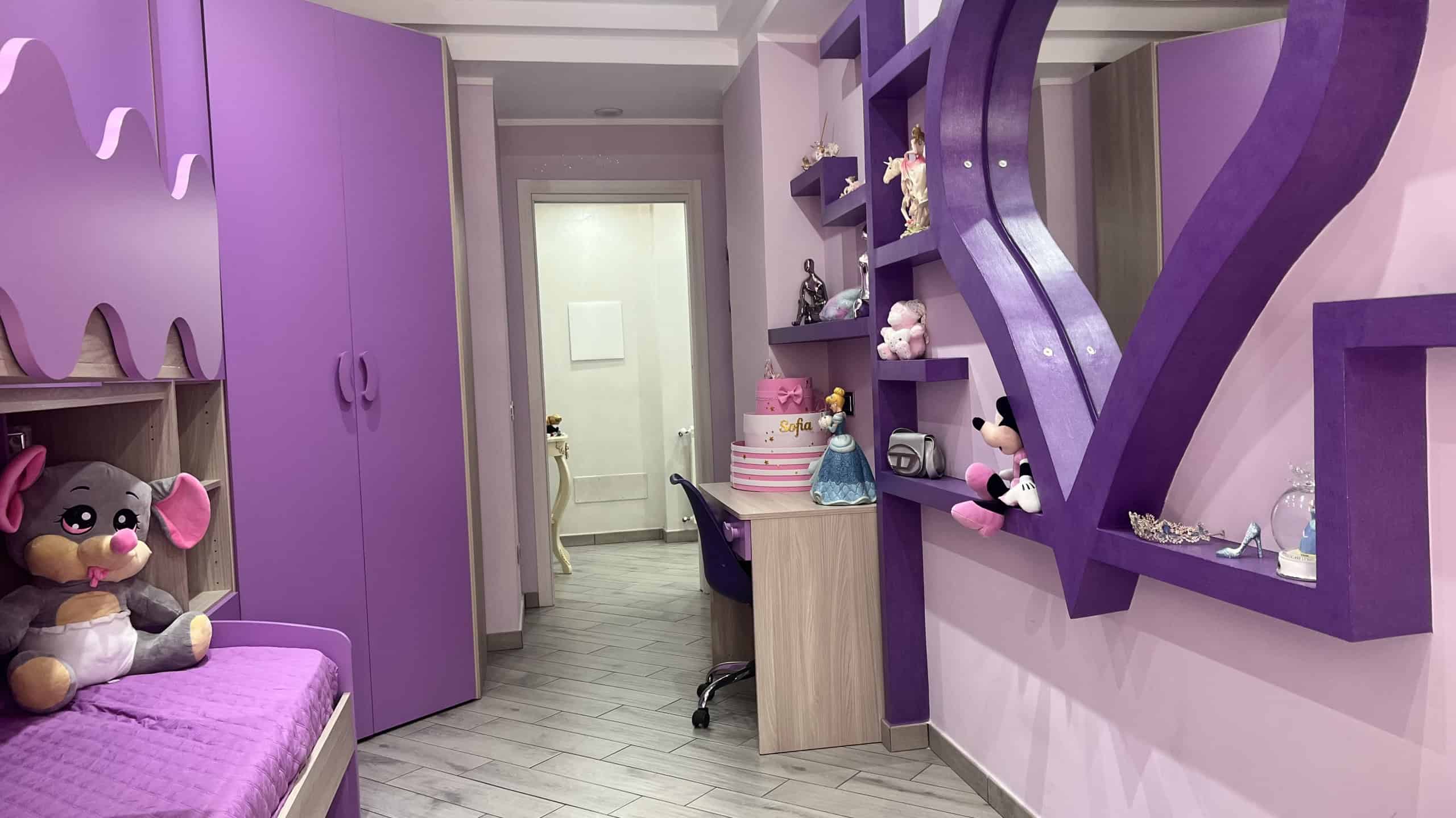 Camera da letto per bambini con arredi viola e decorazioni Disney, con armadio, scrivania e giochi.