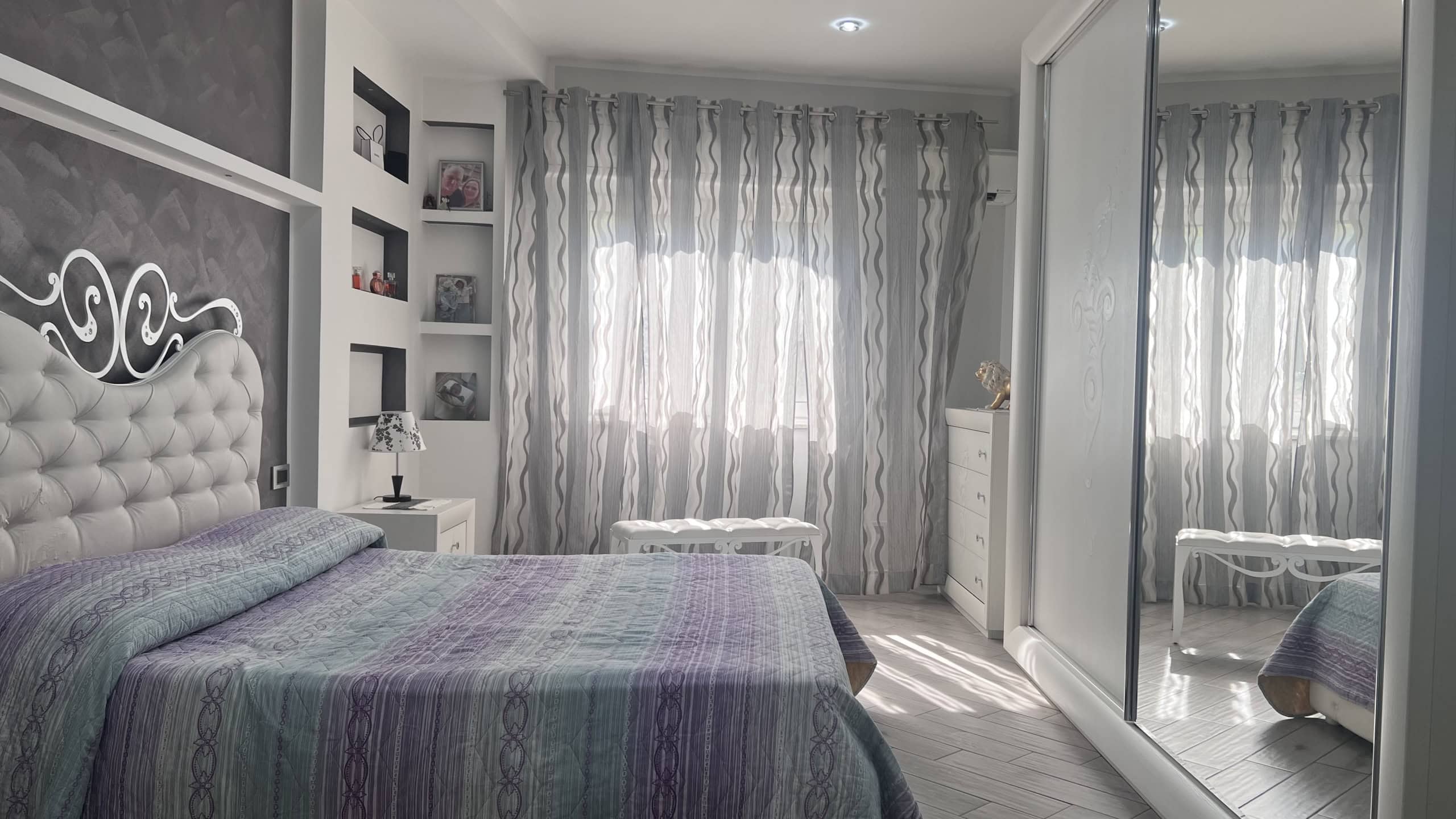 Interno di una camera da letto luminosa con pareti decorate, grande letto con testiera imbottita, armadio a specchio e tende leggere, in un appartamento in vendita a Messina.