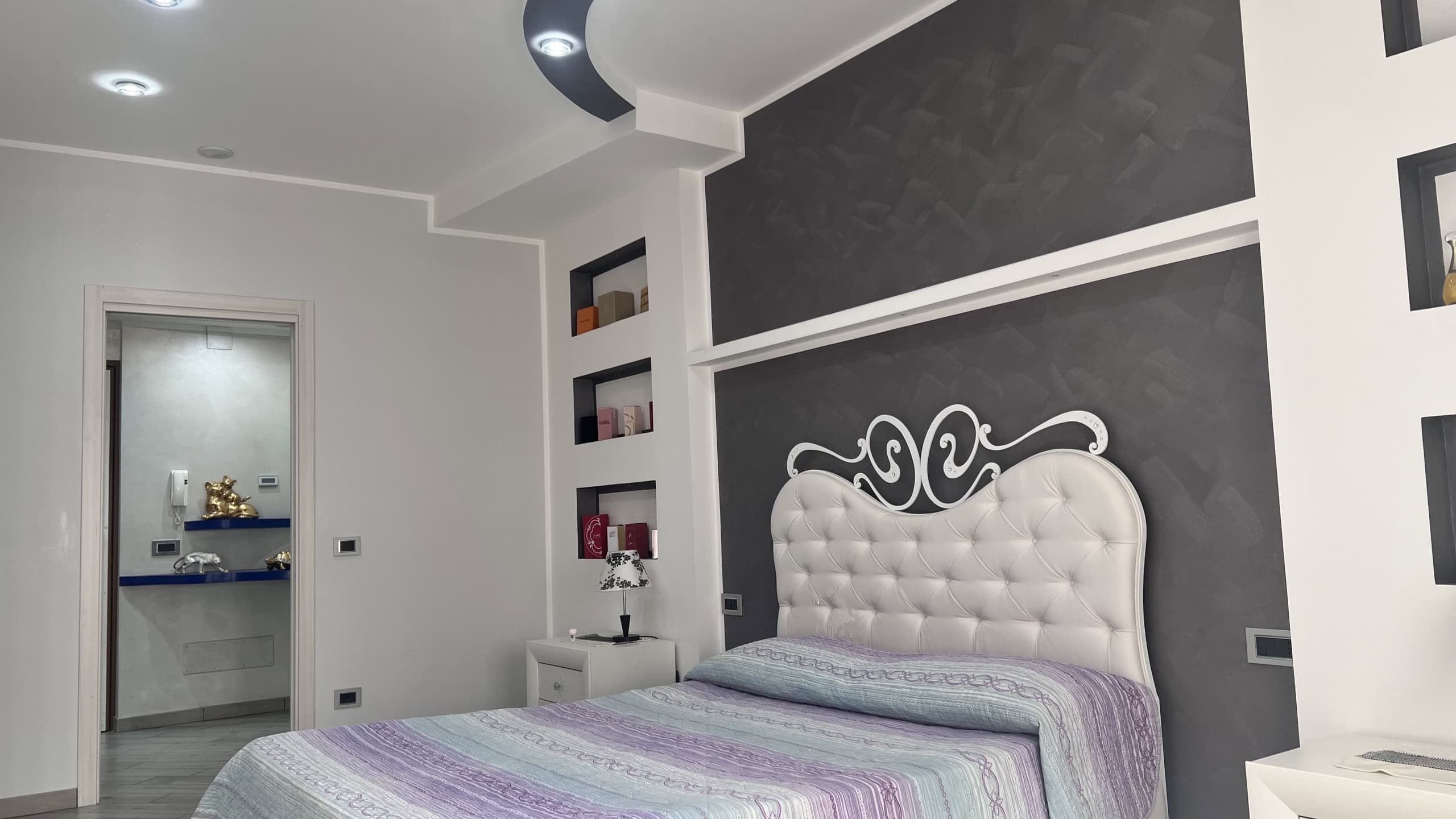 Interno di una camera da letto moderna con pareti bianche e nere, letto con testiera imbottita e decorazioni eleganti, in un appartamento in vendita a Messina.
