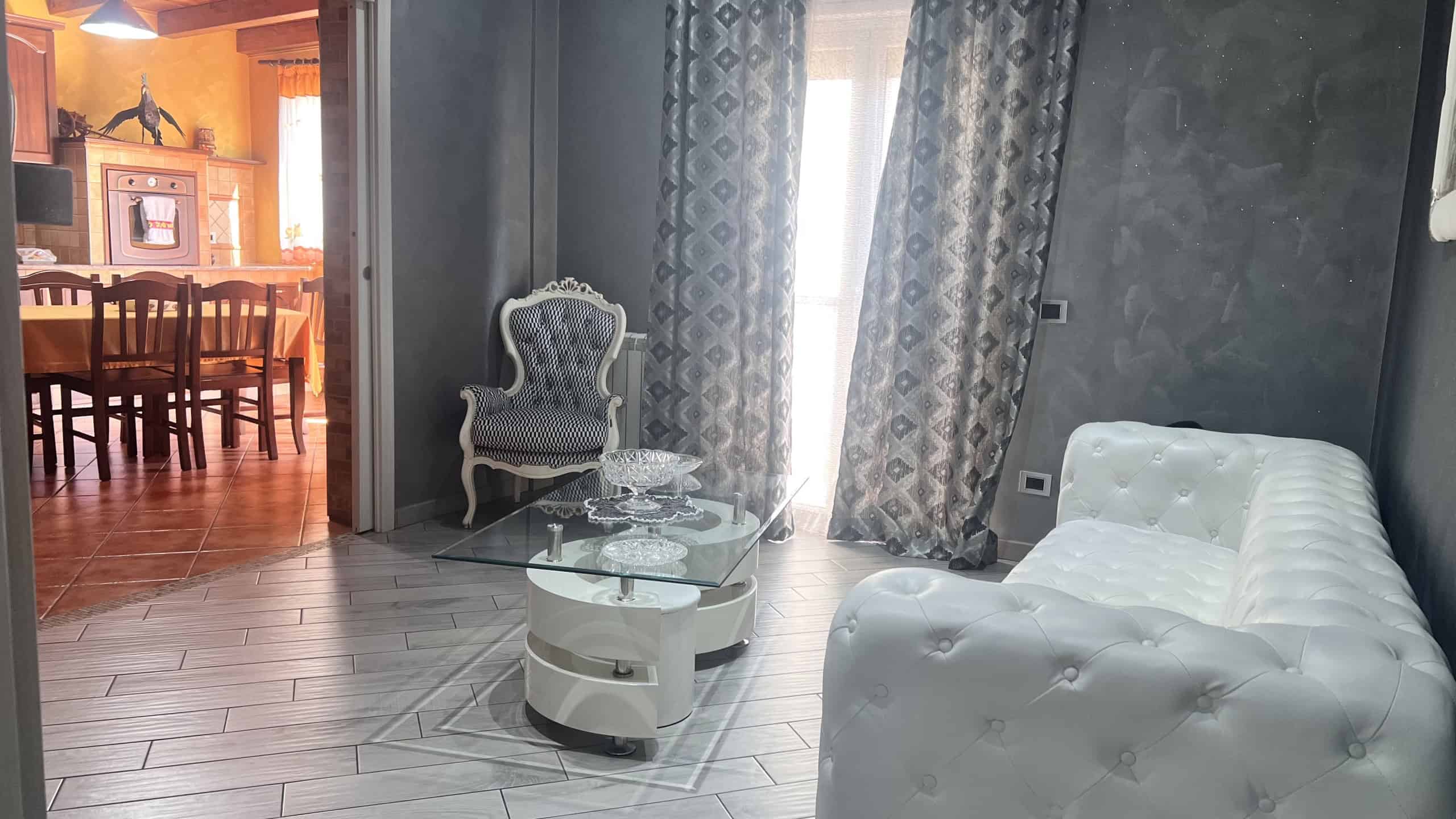 Interno di un salotto elegante con divano bianco, poltrona decorativa e grandi finestre con tende ornate, in un appartamento in vendita a Messina.