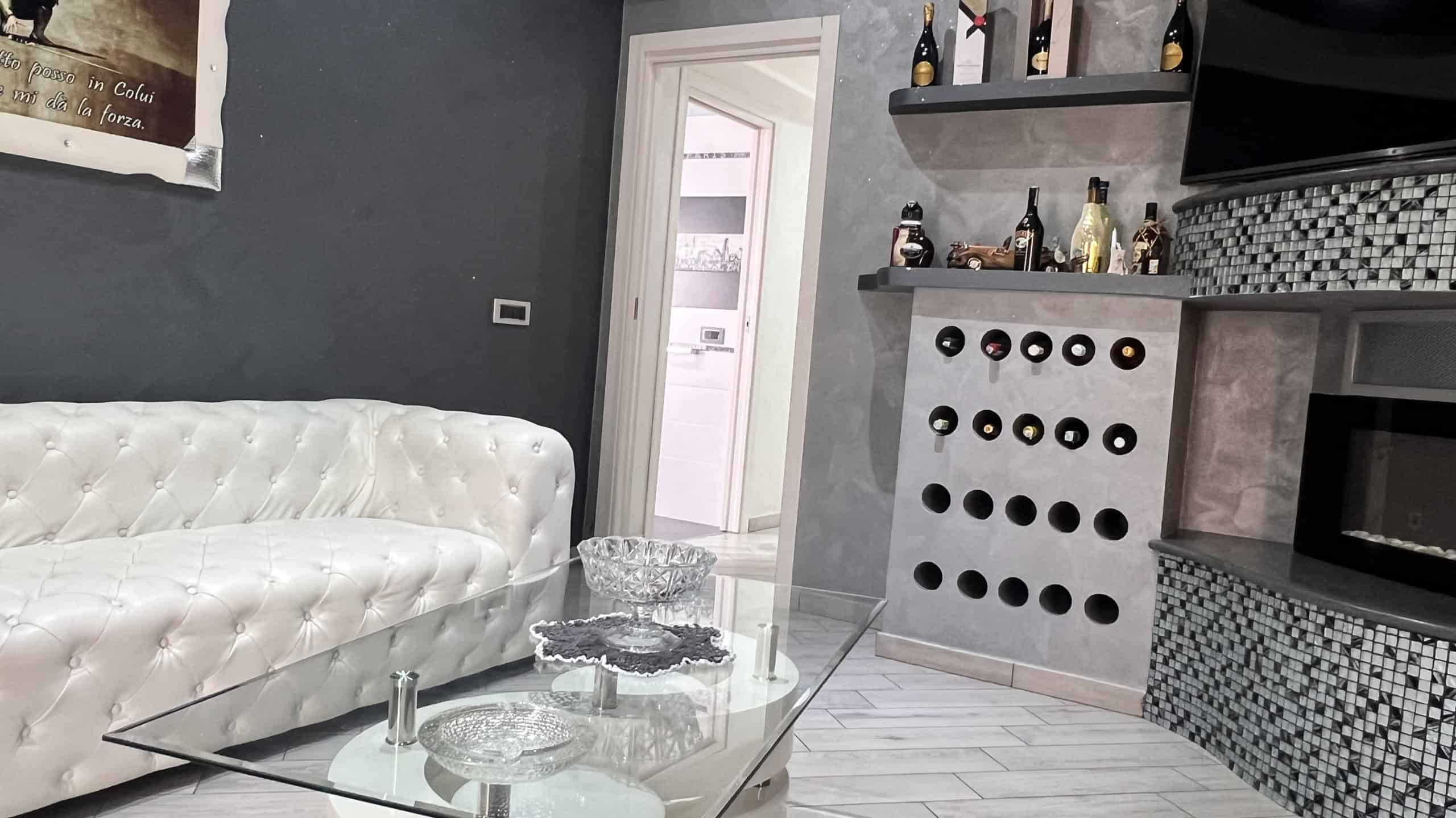 Interno di un soggiorno elegante con divano bianco, tavolino in vetro e pareti grigie, vicino a una cucina con scaffali e bottiglie di vino.