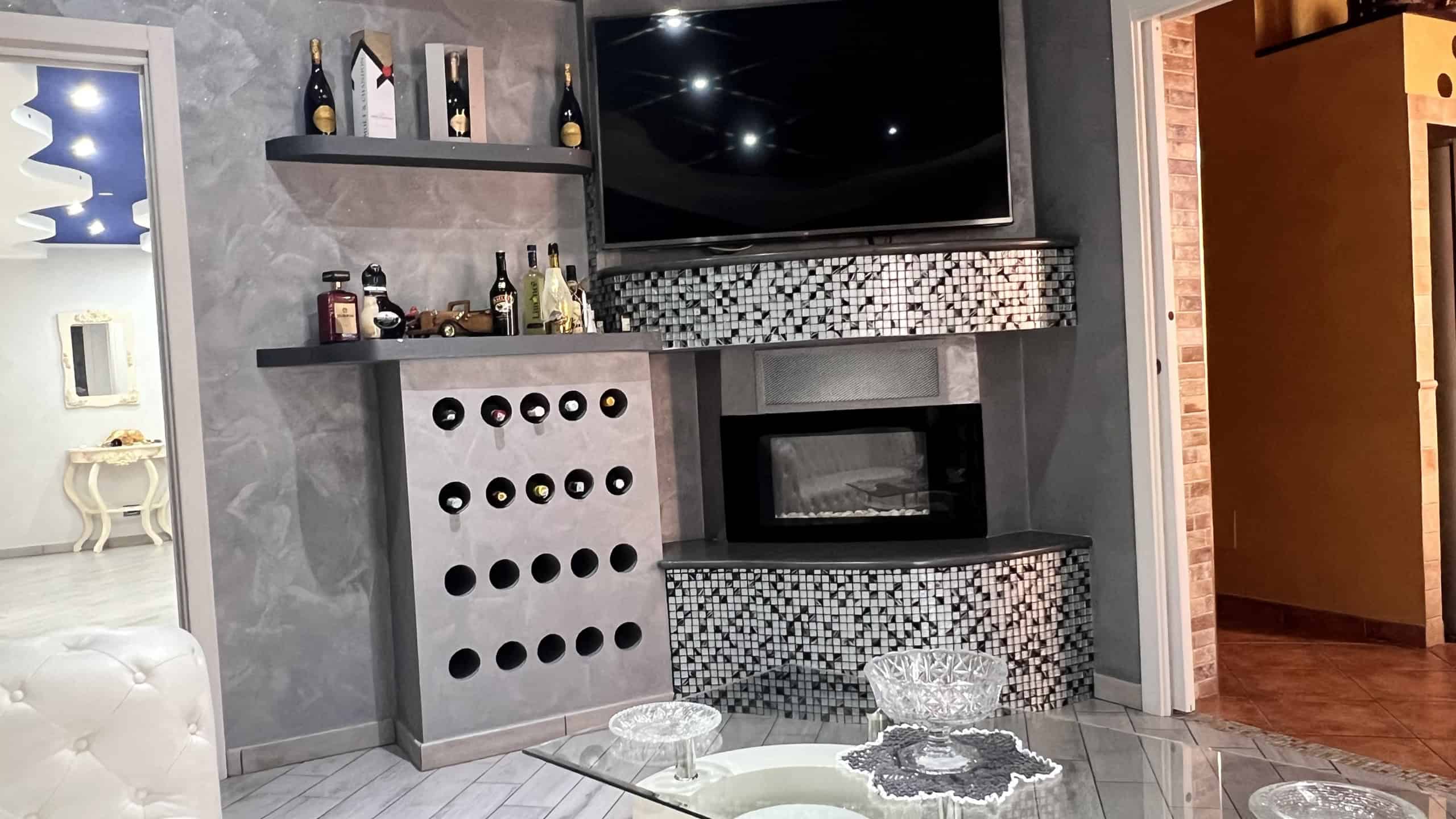 Salotto elegante con camino a gas, TV a schermo piatto e scaffali con bottiglie di vino, arredamento contemporaneo in tonalità grigie e nere.